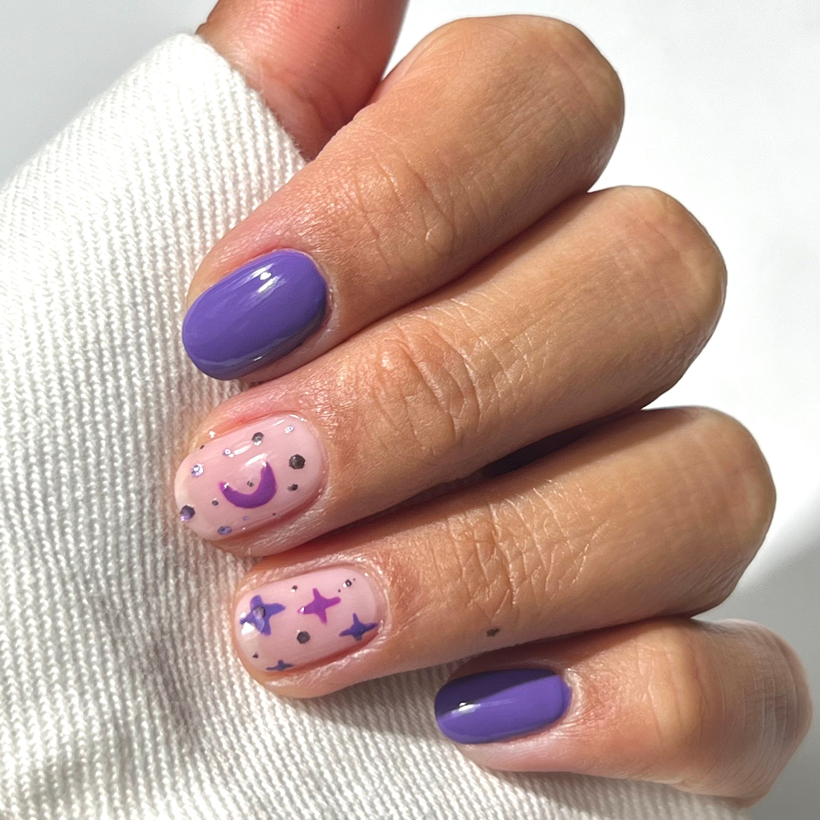A hand with purple and nude nail polish; nude nails feature purple star and moon designs with small dots, while other nails are painted solid purple. The hand rests on a white fabric.