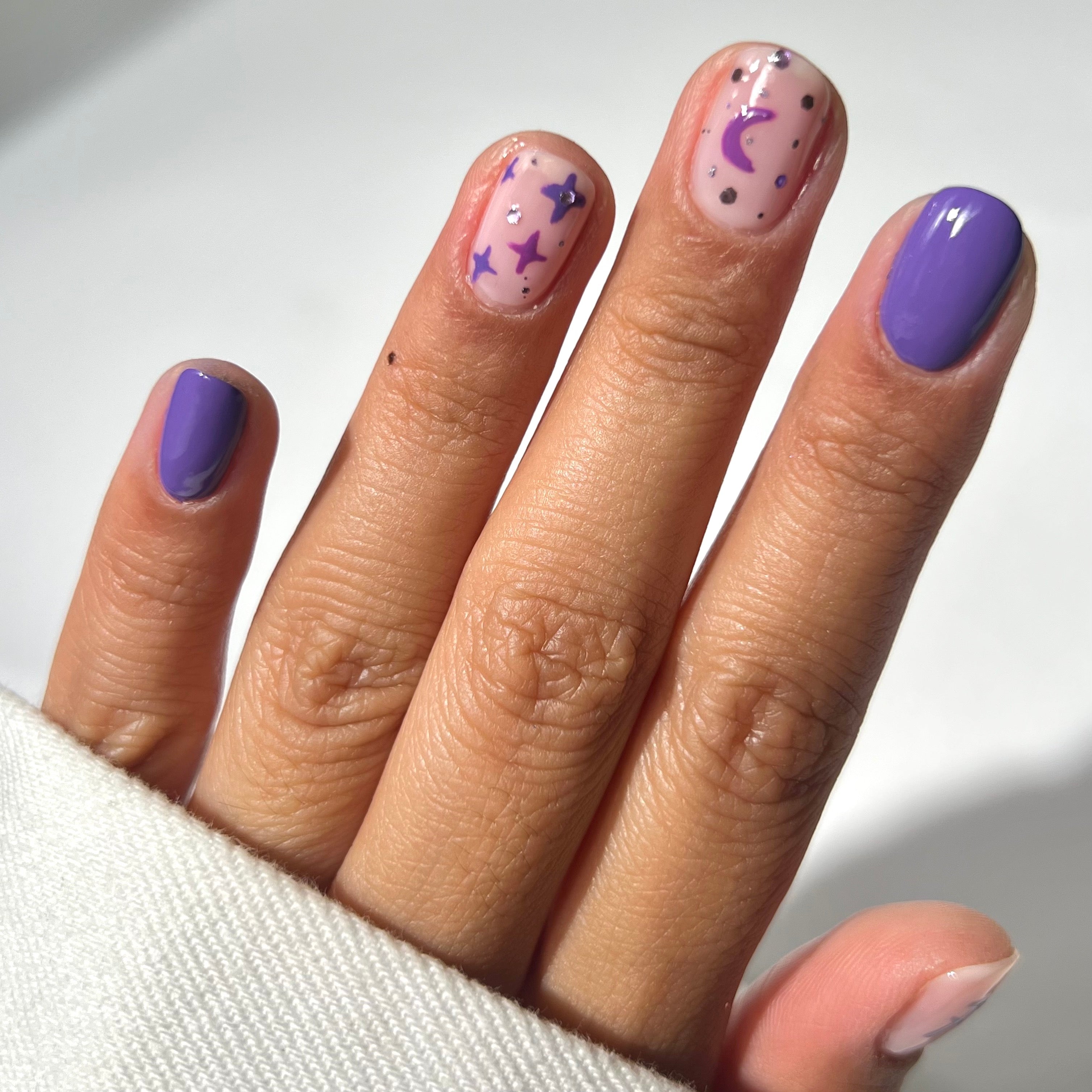 A hand with nails painted in purple and nude colors; two nails have star, moon, and dot designs in purple. The person is wearing a white long-sleeve garment.
