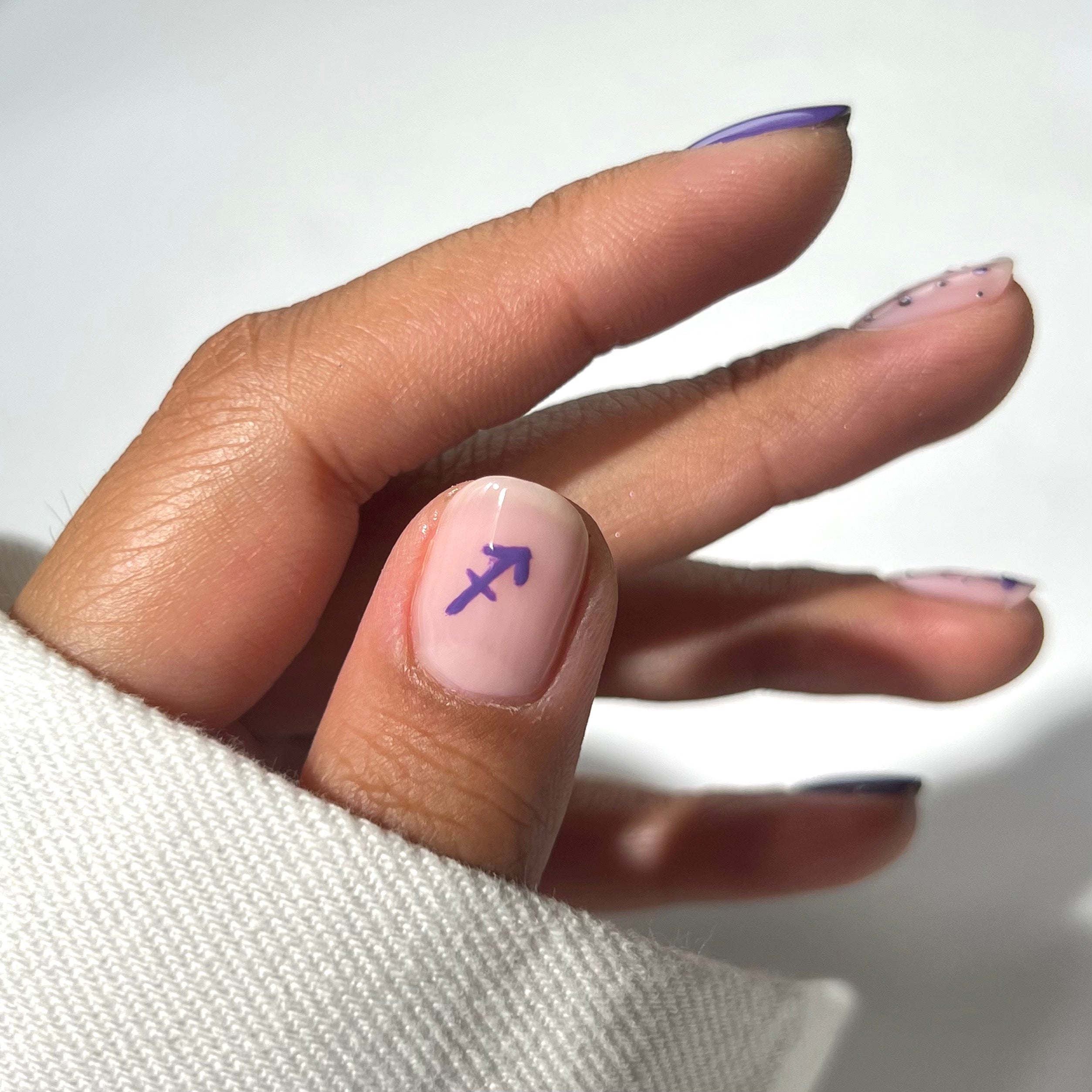 A hand with manicured nails; the thumb features a purple Sagittarius zodiac symbol on a nude base, while the other nails have subtle purple nail art, all peeking out from a white sleeve.