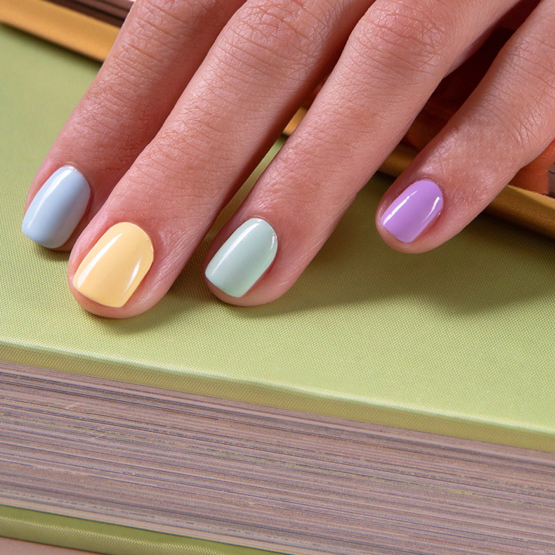 A hand with pastel-colored nail polish in blue, yellow, green, and purple rests on a closed, light green book.
