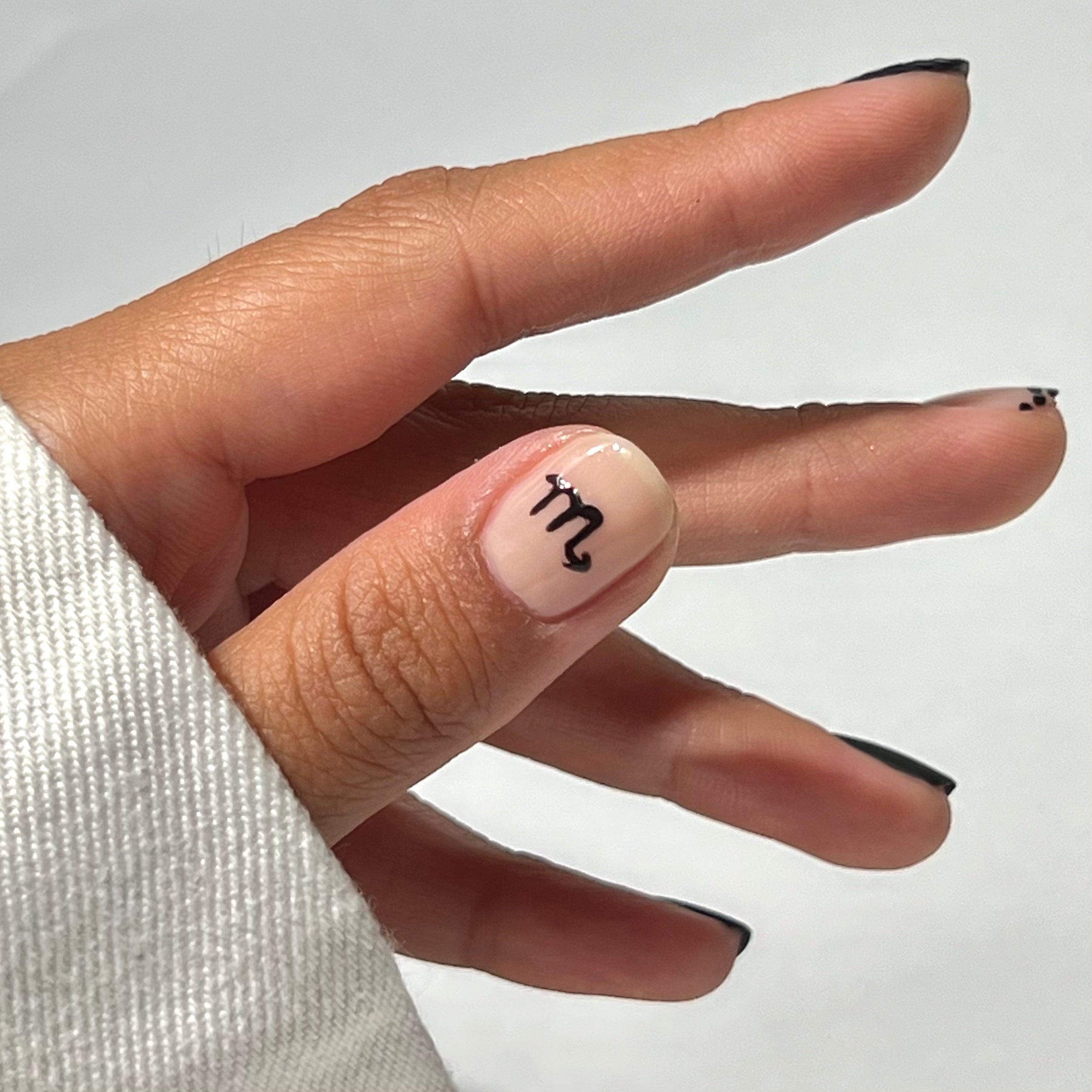 A hand with black-tipped nails and a Scorpio zodiac symbol painted on the thumbnail. The person is wearing a white long-sleeve shirt. The background is plain and light-colored.