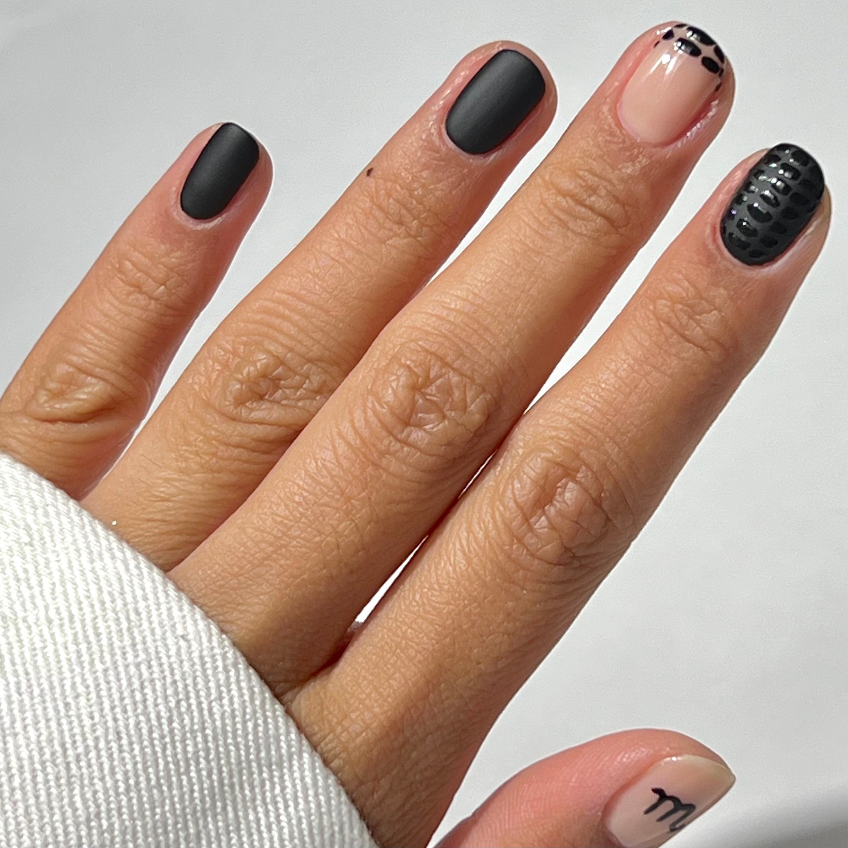 A hand with a manicure featuring matte black nails, one nude nail with black polka dots, one nude nail with a small black design near the cuticle, and a thumb with black lettering.
