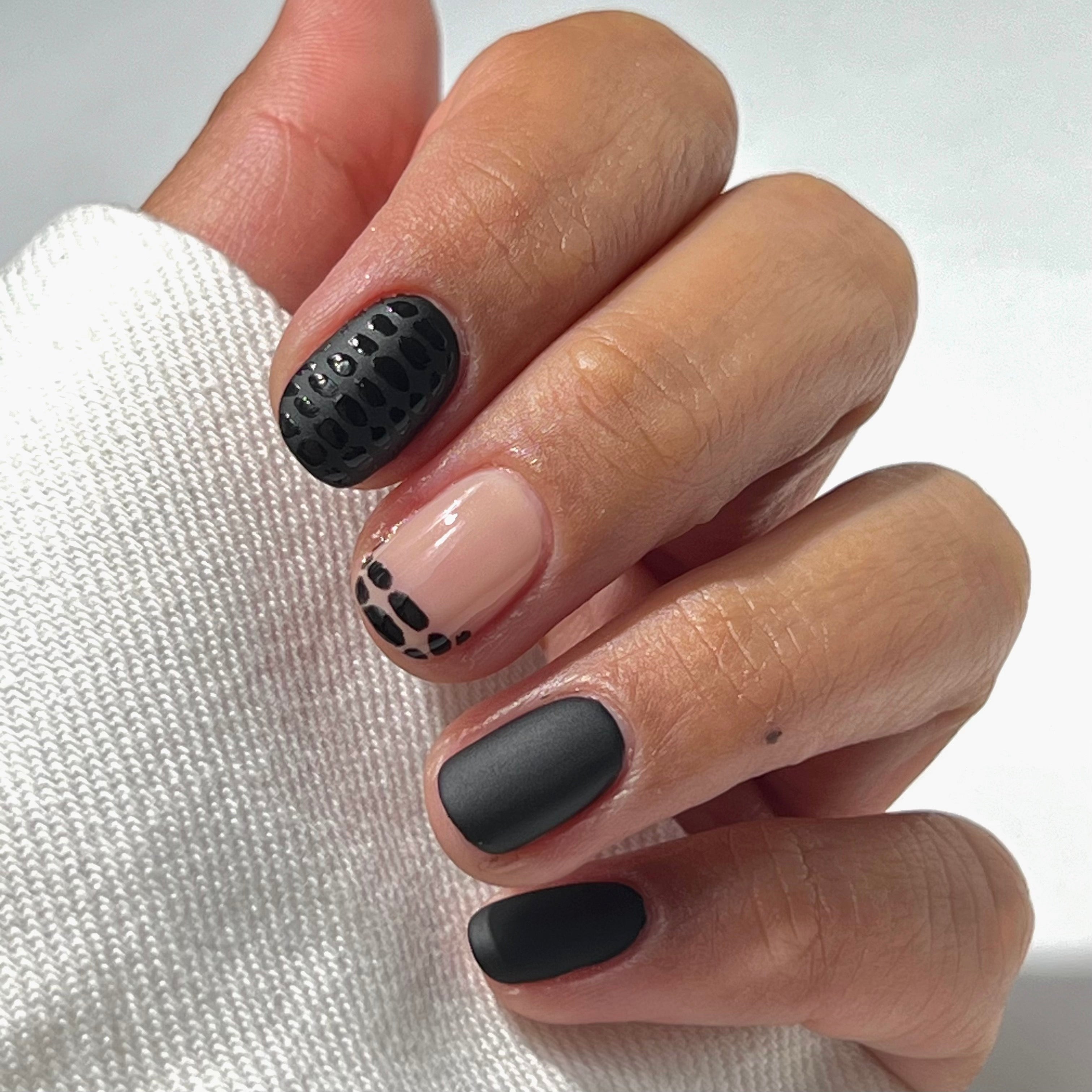 A hand with short, manicured nails featuring black and nude nail art: one nail has black scales, one is matte black, one is nude with small black dots, and another is glossy black. The thumb is partially visible in white fabric.