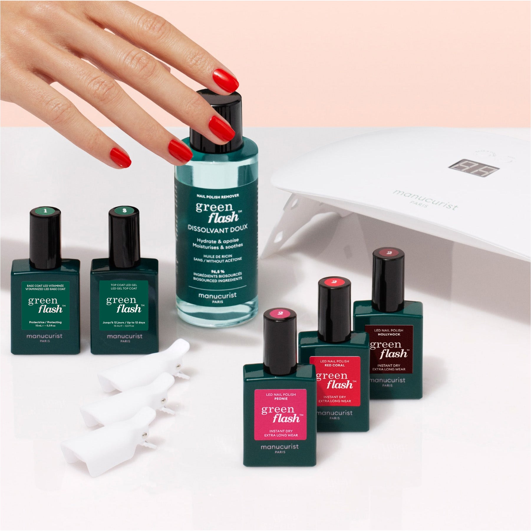 A hand with red-painted nails reaches for a Manucurist Starter Kit, featuring nail polish remover, gel polishes, fingertip clips, and an LED lamp—everything needed for a salon-quality manicure at home.