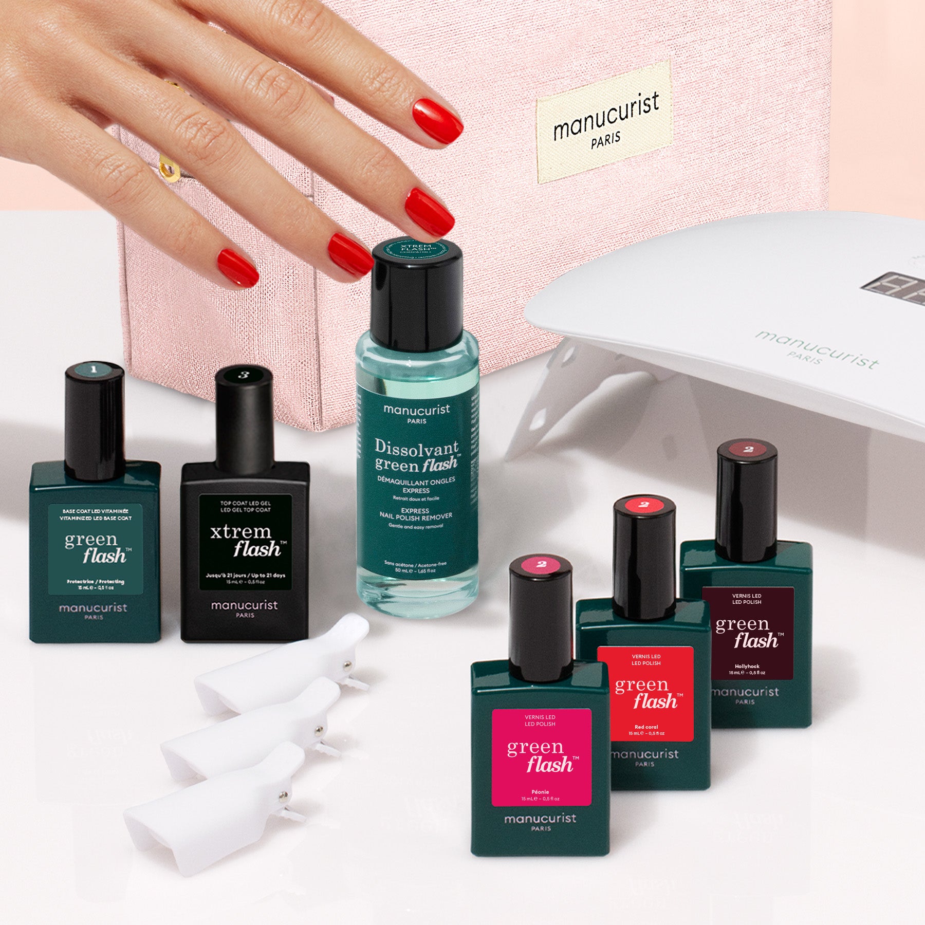 A hand with red nail polish is surrounded by the Manucurist Starter Kit, which includes nail polish bottles, a vitamin-infused base coat, remover, nail clips, a pink pouch, and an LED nail lamp—everything you need for an ultra-glossy manicure.
