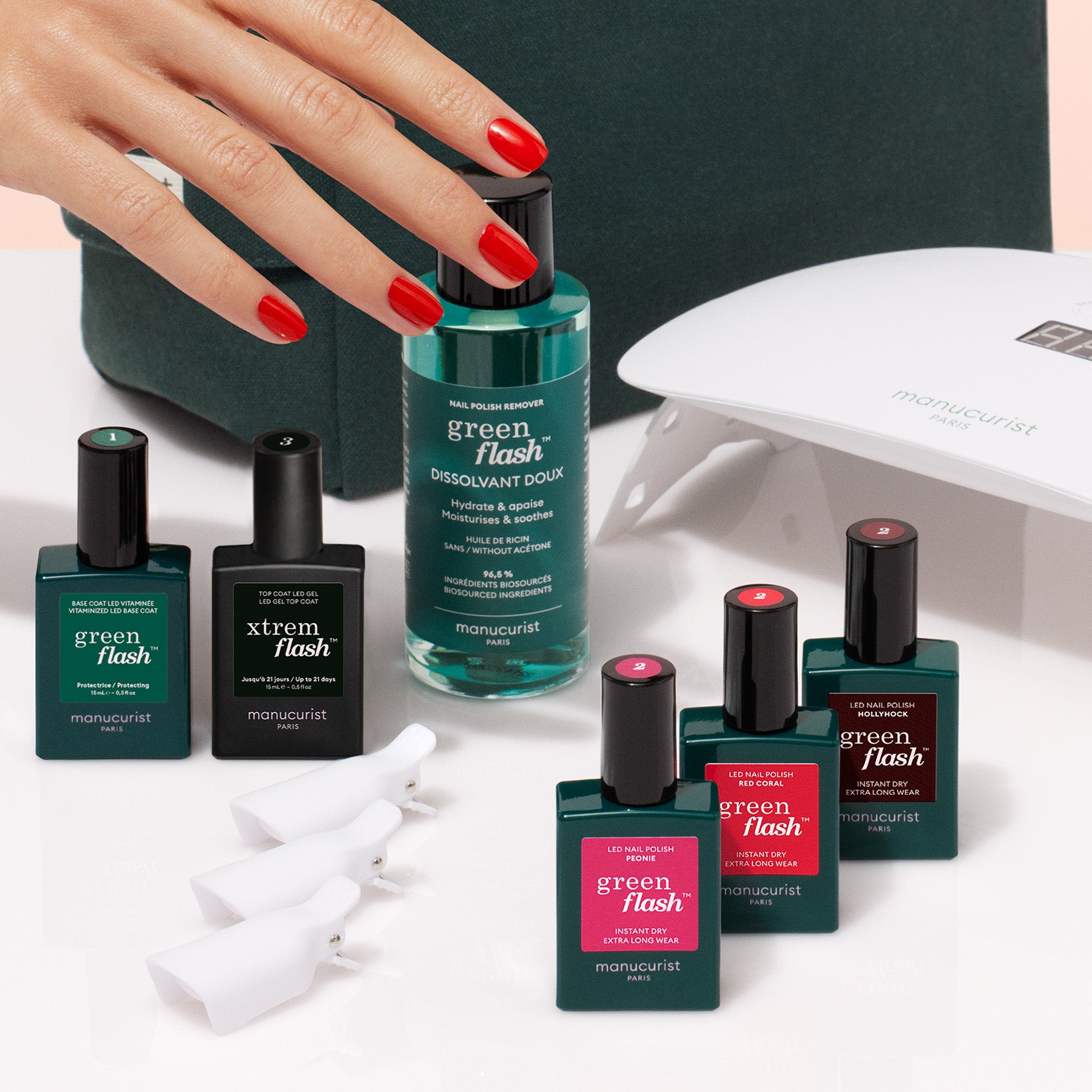 A hand with red-painted nails rests near Manucurist’s Starter Kit, featuring a remover bottle, white nail lamp, and white clips—must-haves for an ultra-glossy manicure—neatly arranged on a white surface.