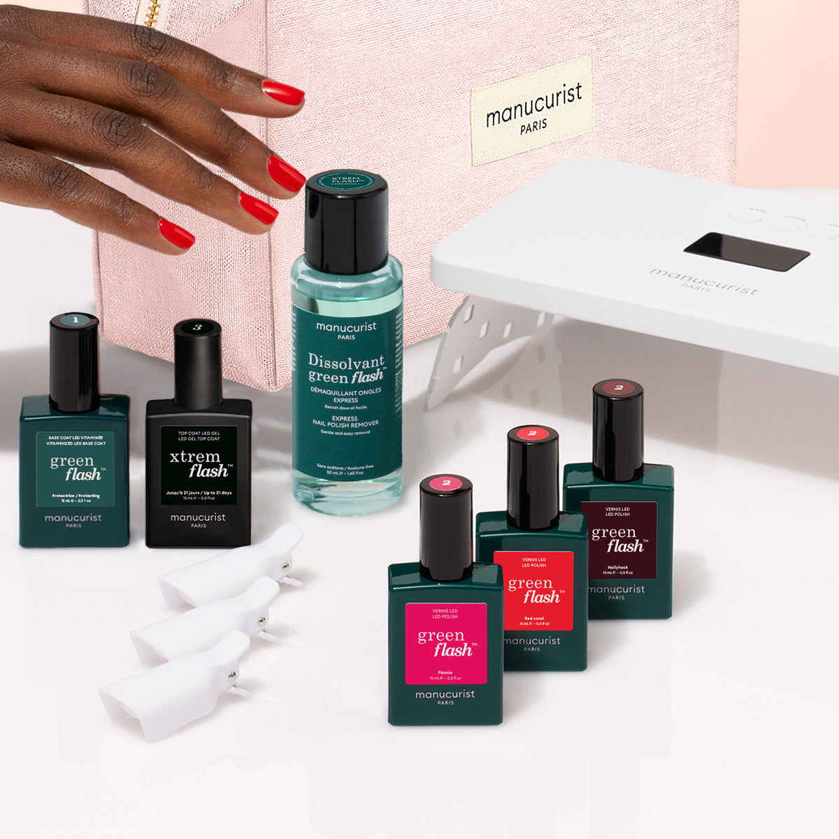 A hand reaches for the Manucurist Starter Kit—featuring a pink gel polish—next to bottles of Manucurist nail polish, a vitaminized base coat, dissolvant, nail clips, and an LED lamp on a white surface.