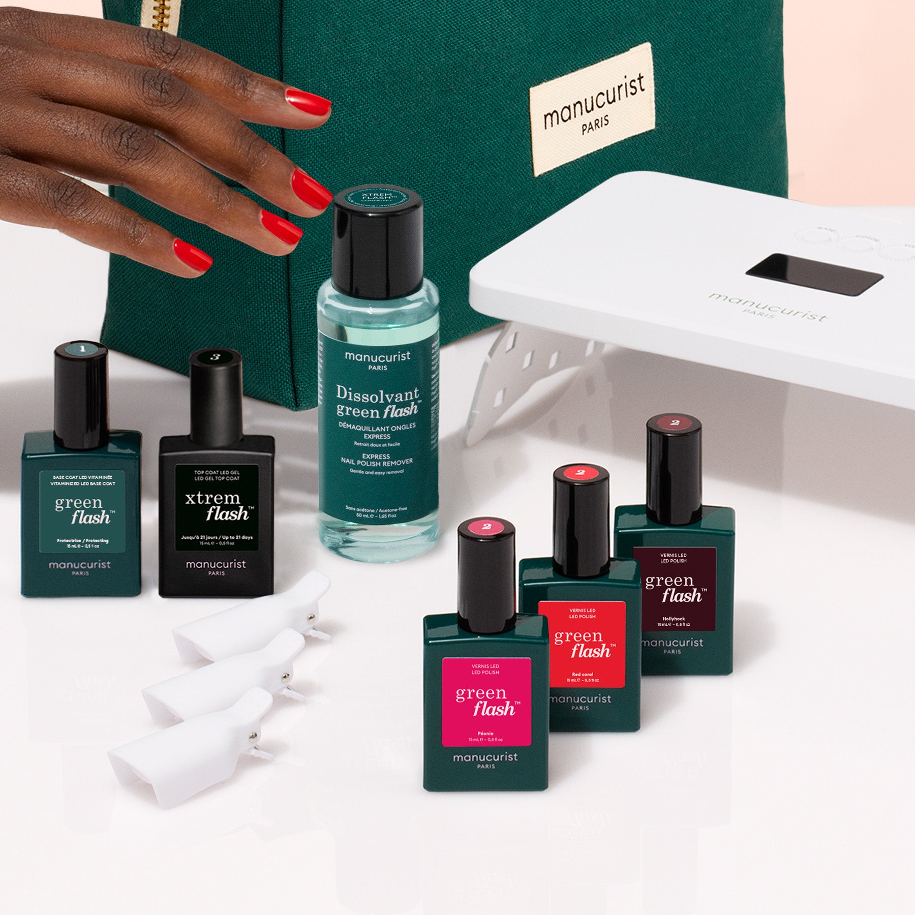 A hand with red nails reaches for the Manucurist Starter Kit, featuring all essentials for an ultra-glossy manicure—vitaminized base coat, polish remover, clips, UV lamp, and a green Paris pouch—arranged on a white surface.