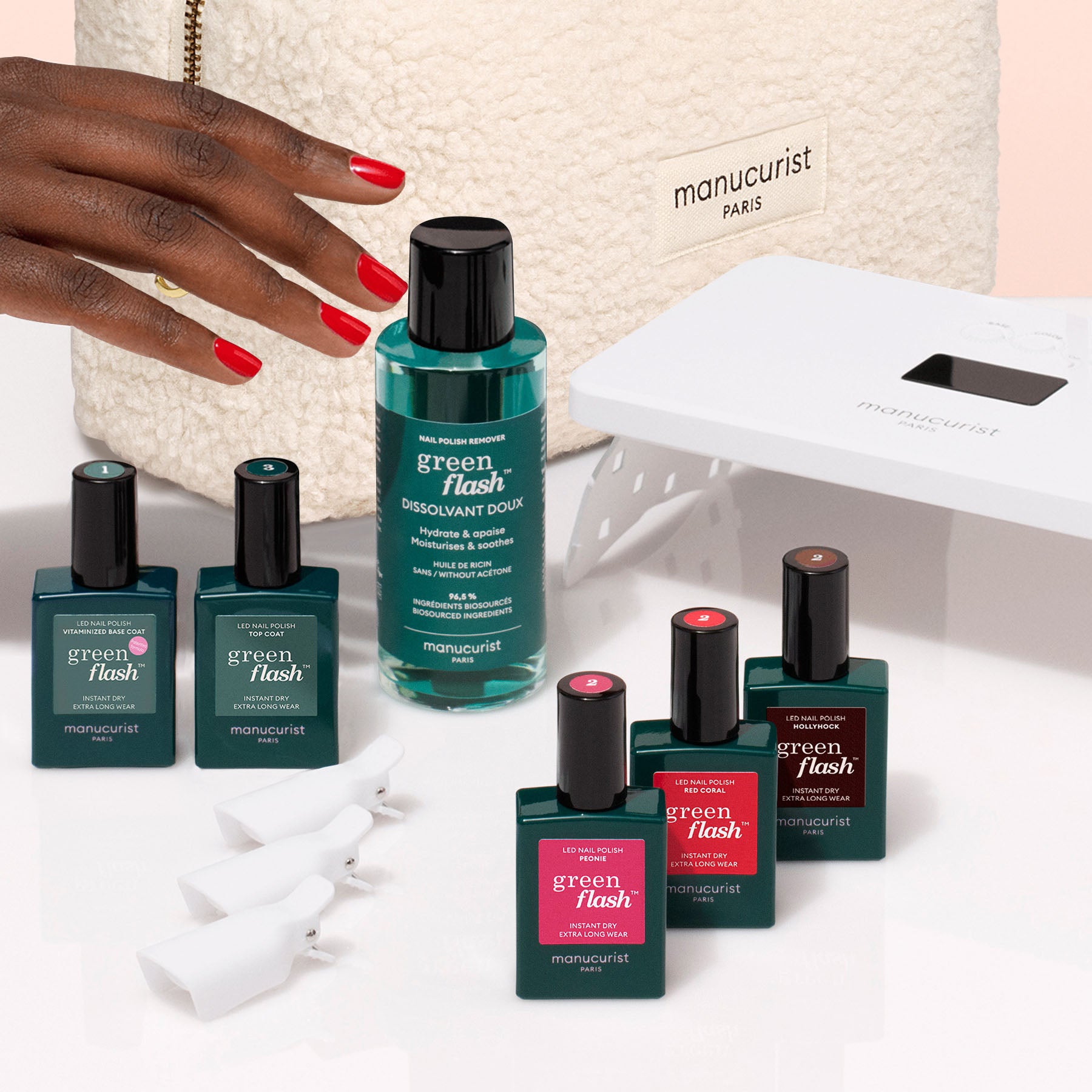 A hand with red nails reaches for the Manucurist Starter Kit, which includes nail polishes, remover, nail clips, a vitaminized base coat, UV lamp, and a cream-colored pouch for an ultra-glossy manicure.