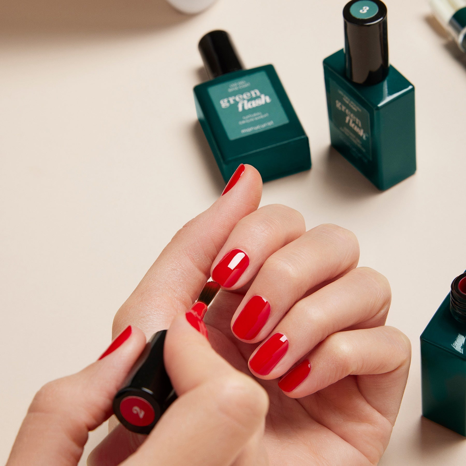 Someone is applying red gel polish from the Manucurist-test Green Flash™ Welcome Kit to their nails, with green-labeled bottles, manicure tools, and acetone-free remover arranged on a light-colored surface nearby.