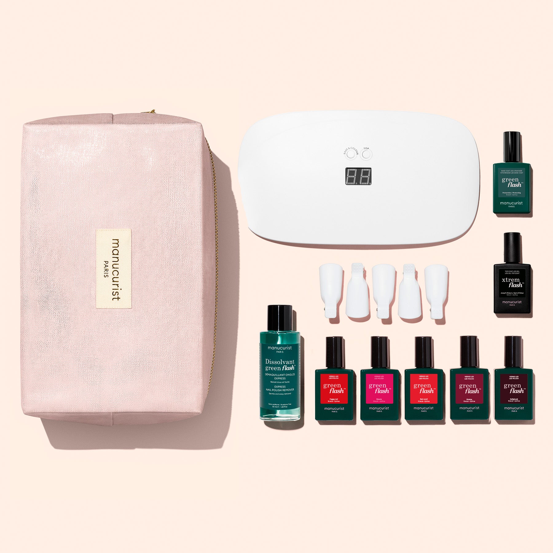 A hand with red nails rests near bottles from the Manucurist Super Starter Kit, including gel polishes, nail polish remover, a UV lamp, removal clips, and a pink bag. The products are arranged on a white surface, promising an ultra-glossy manicure that lasts.
