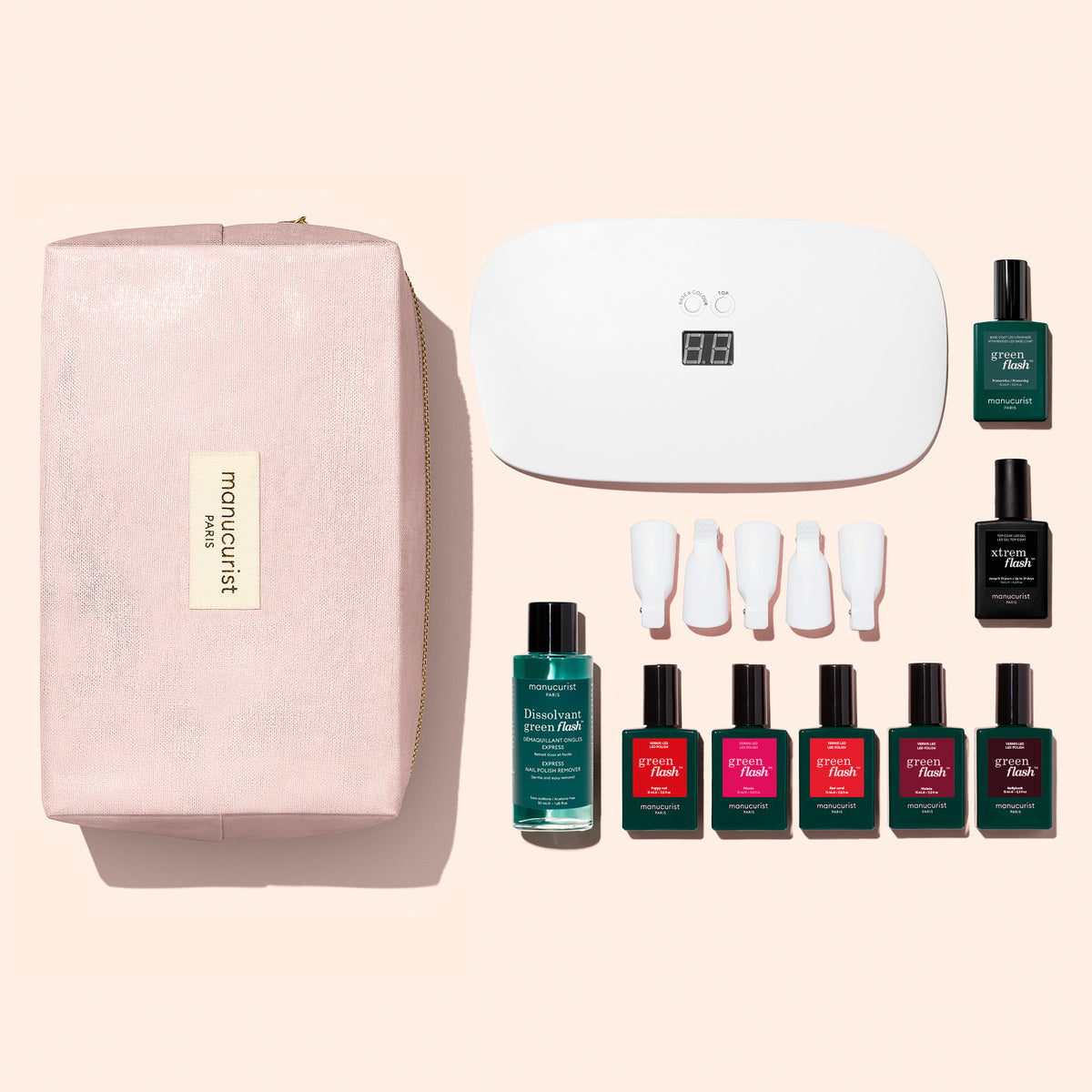 Une main aux ongles rouges est posée près des flacons du Super Starter Kit Manucurist, comprenant des vernis gel, du dissolvant, une lampe UV, des pinces de retrait et une trousse rose. Les produits sont disposés sur une surface blanche, promettant une manucure ultra-brillante qui dure.