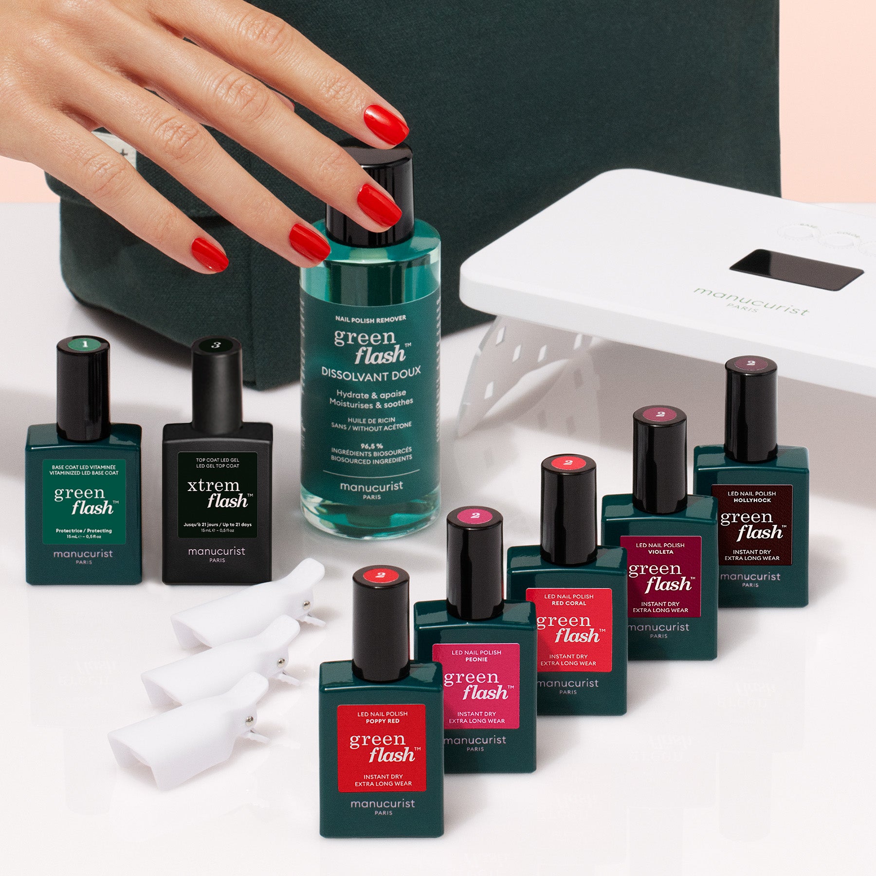 A hand with red nails reaches for the Manucurist Super Starter Kit, featuring gel polish, remover, manicure tools, a dark case, and a nail lamp on a white surface for an ultra-glossy look.