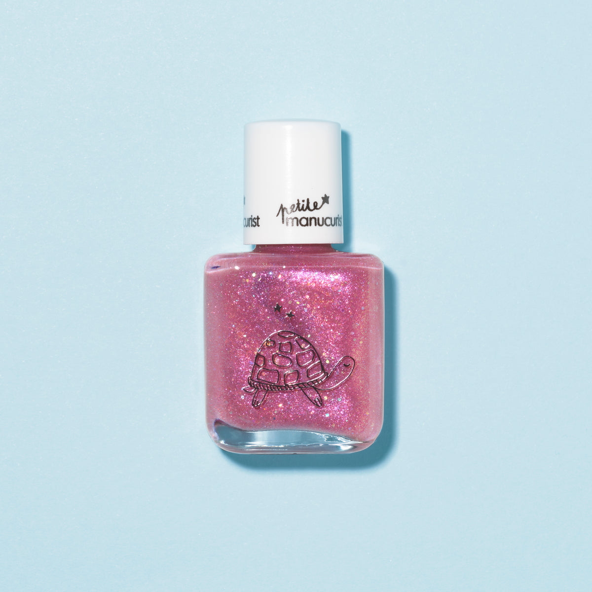 A clear bottle of Manucurist’s “Betty the Turtle” raspberry glitter pink kids’ nail polish with a white cap and turtle illustration, set on a light blue background. Easily removable with soapy water for simple cleanup.
