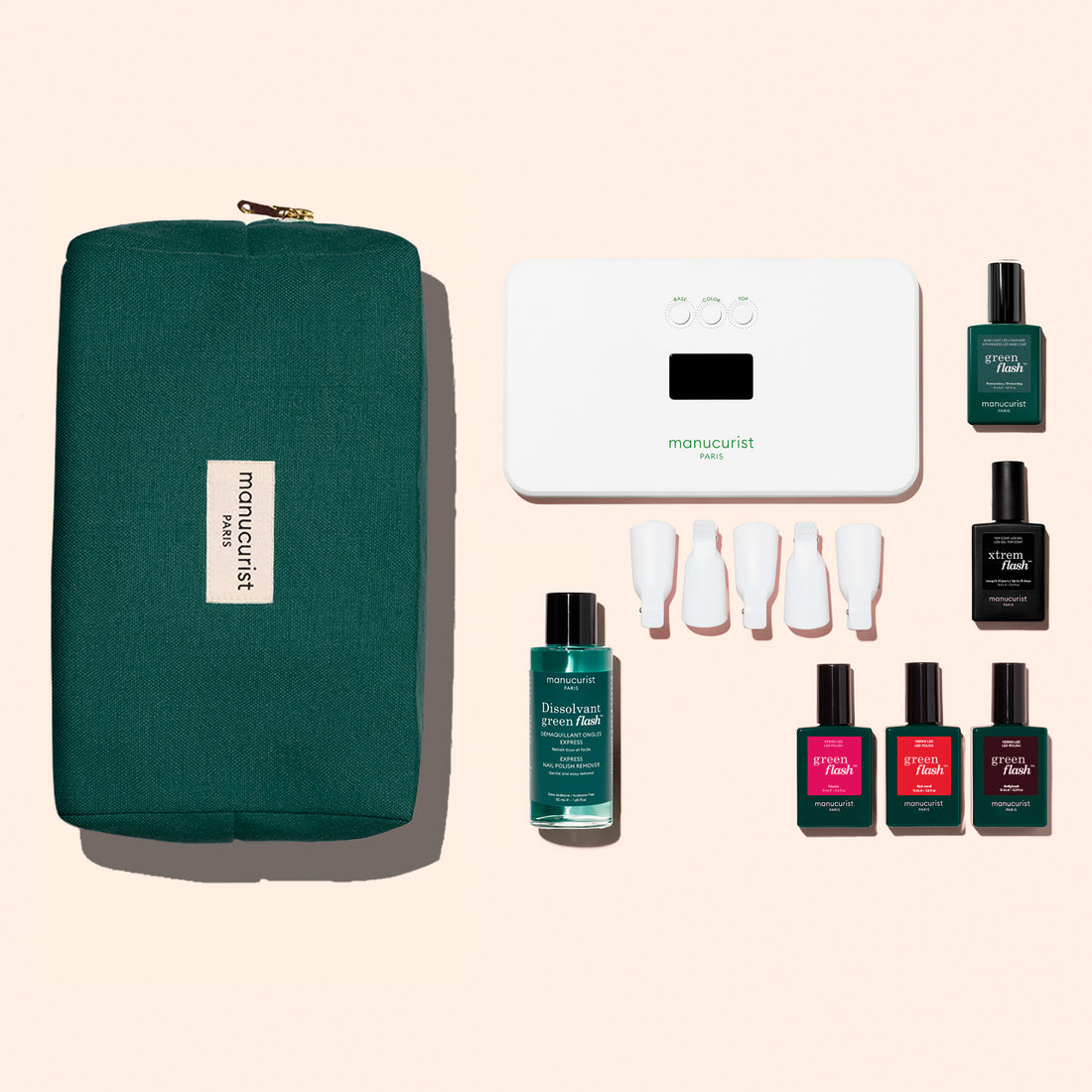 The Manucurist Starter Kit, featuring a green zippered pouch, LED nail lamp, base coat, remover, four caps, and six colored polishes, is neatly arranged on a light pink background.