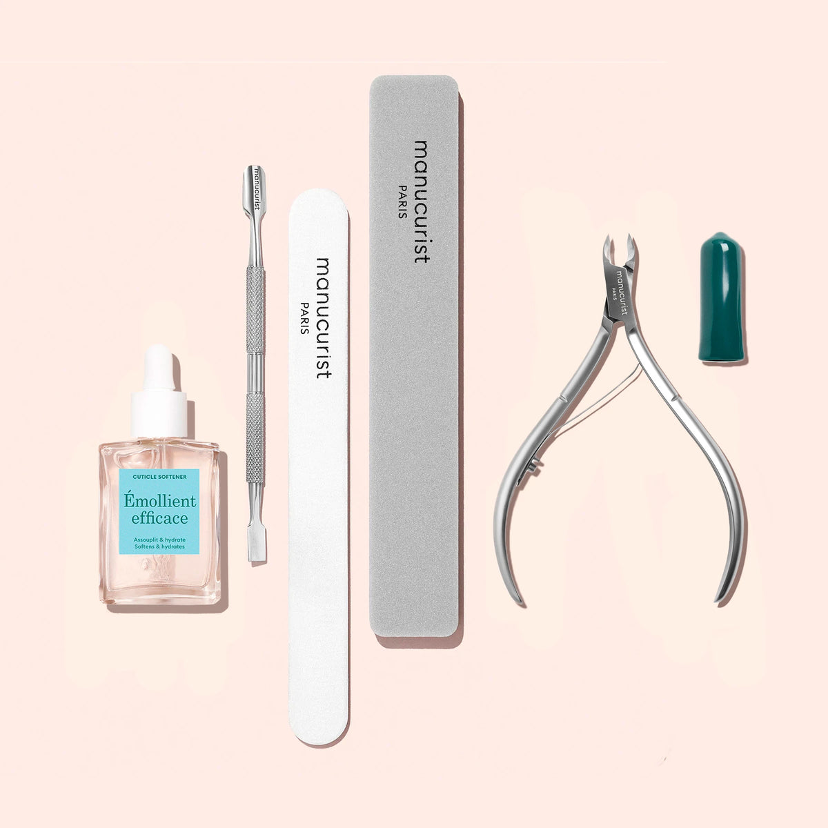 Le Kit Préparation Ongles Manucurist comprend une huile émolliente pour cuticules, un repousse-cuticules en métal, une lime à ongles émeri, un polissoir et une pince à cuticules en acier inoxydable avec capuchon rouge, présentés sur fond rose clair.