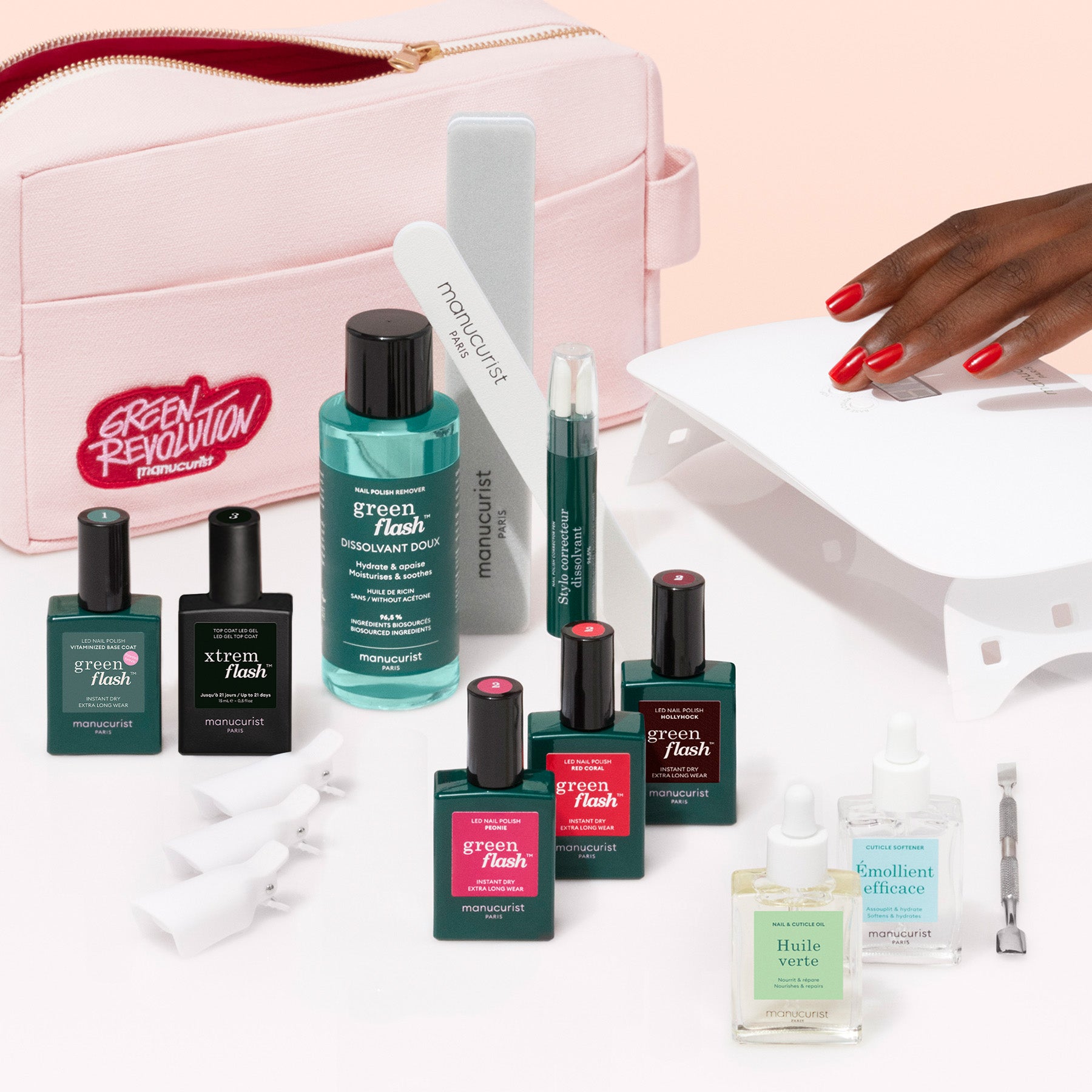 The Manucurist Ultimate Kit, featuring gel polish, Vitaminized Base Coat, remover, oils, nail file, cuticle tool, and LED lamp in a pink cosmetic bag with a Green Revolution patch, is displayed beside a hand with an ultra-glossy manicure.