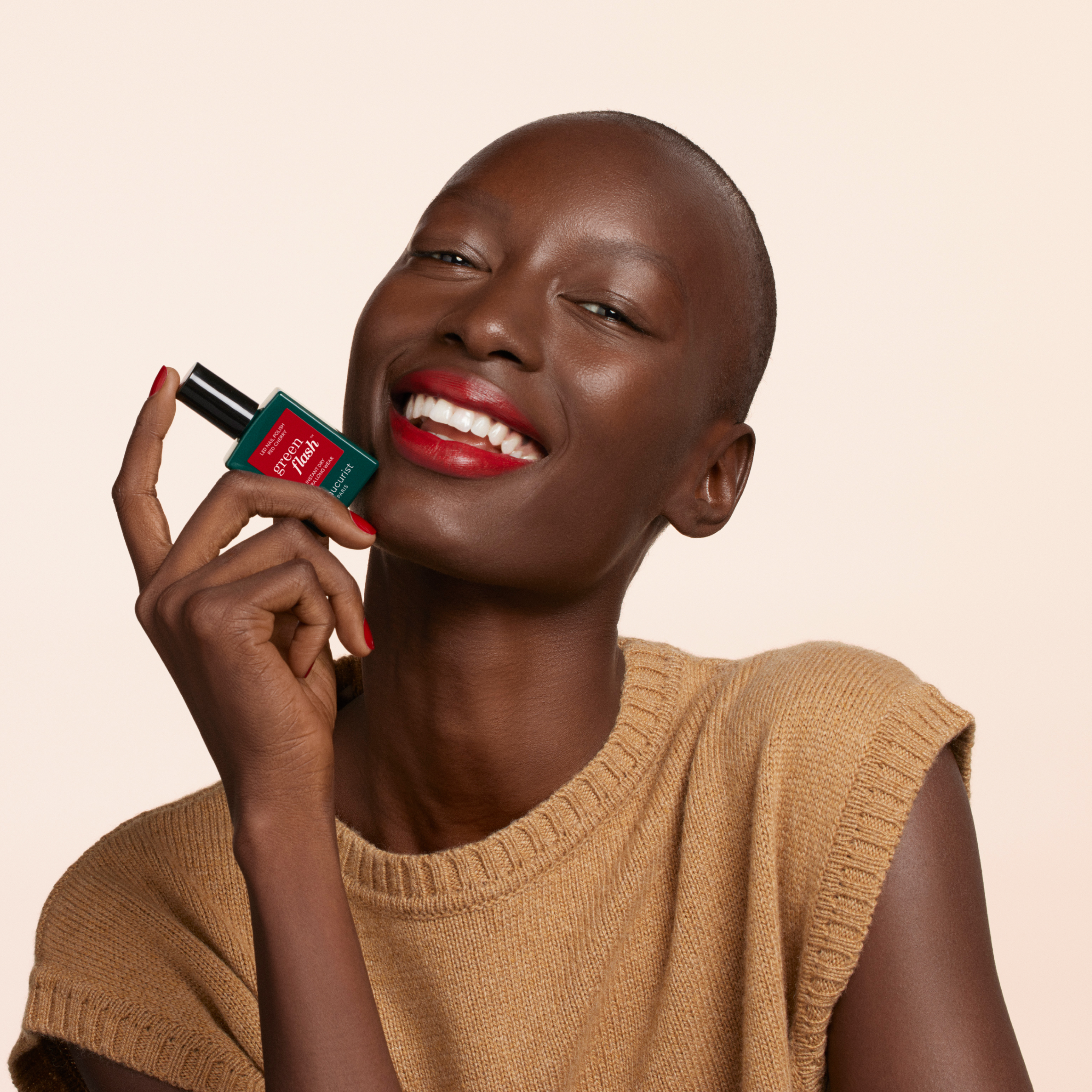 A smiling person with short hair and bright red lipstick holds a green and red perfume bottle near their face, wearing a sleeveless beige knit top against a neutral background.