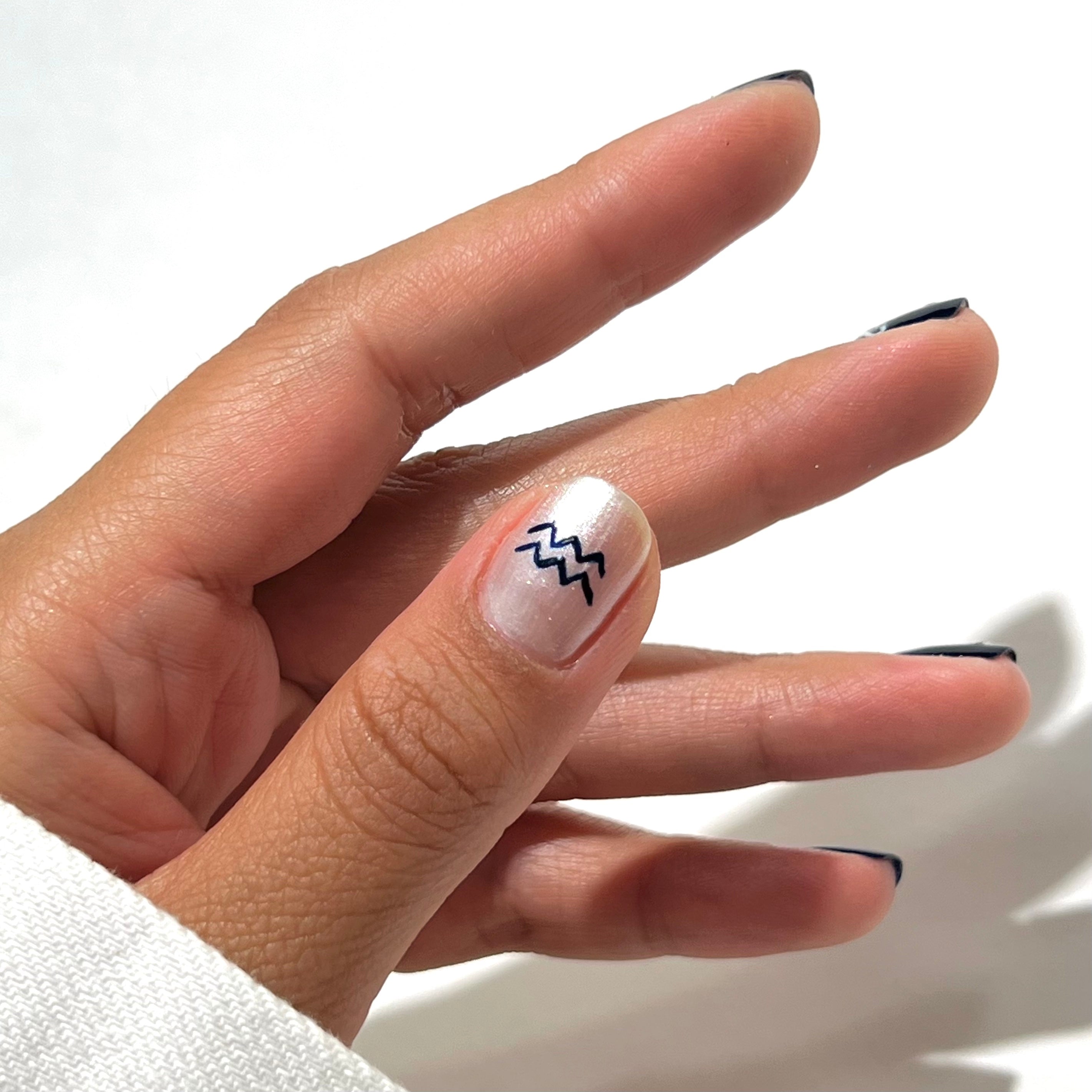 A hand with short, almond-shaped nails. The nails are painted with a sheer nude polish and have thin black tips. The thumb features a small black zigzag line design on a white base.
