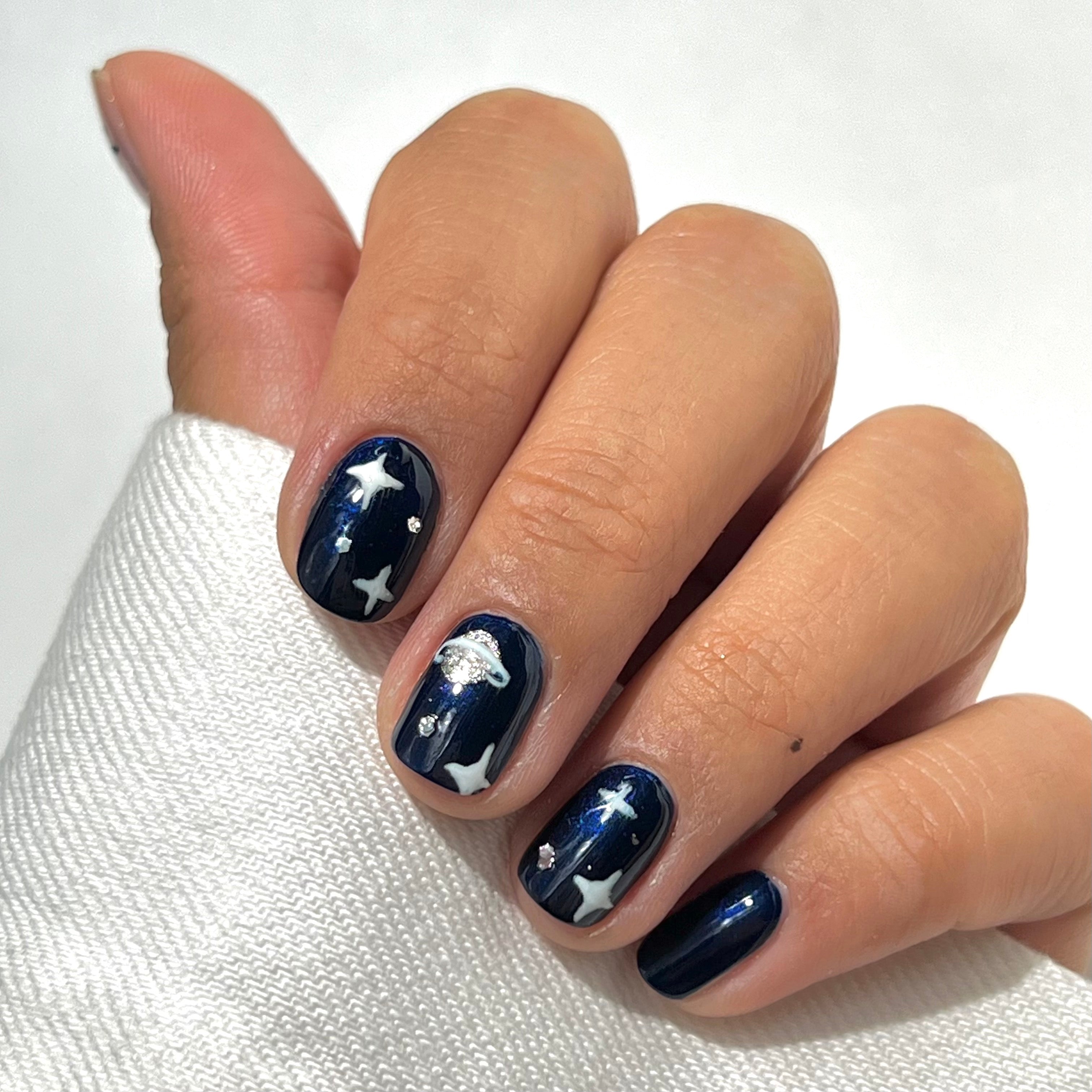 A hand with short, neatly manicured nails painted dark blue, featuring white designs of clouds and stars. The person is wearing a white long-sleeve top.