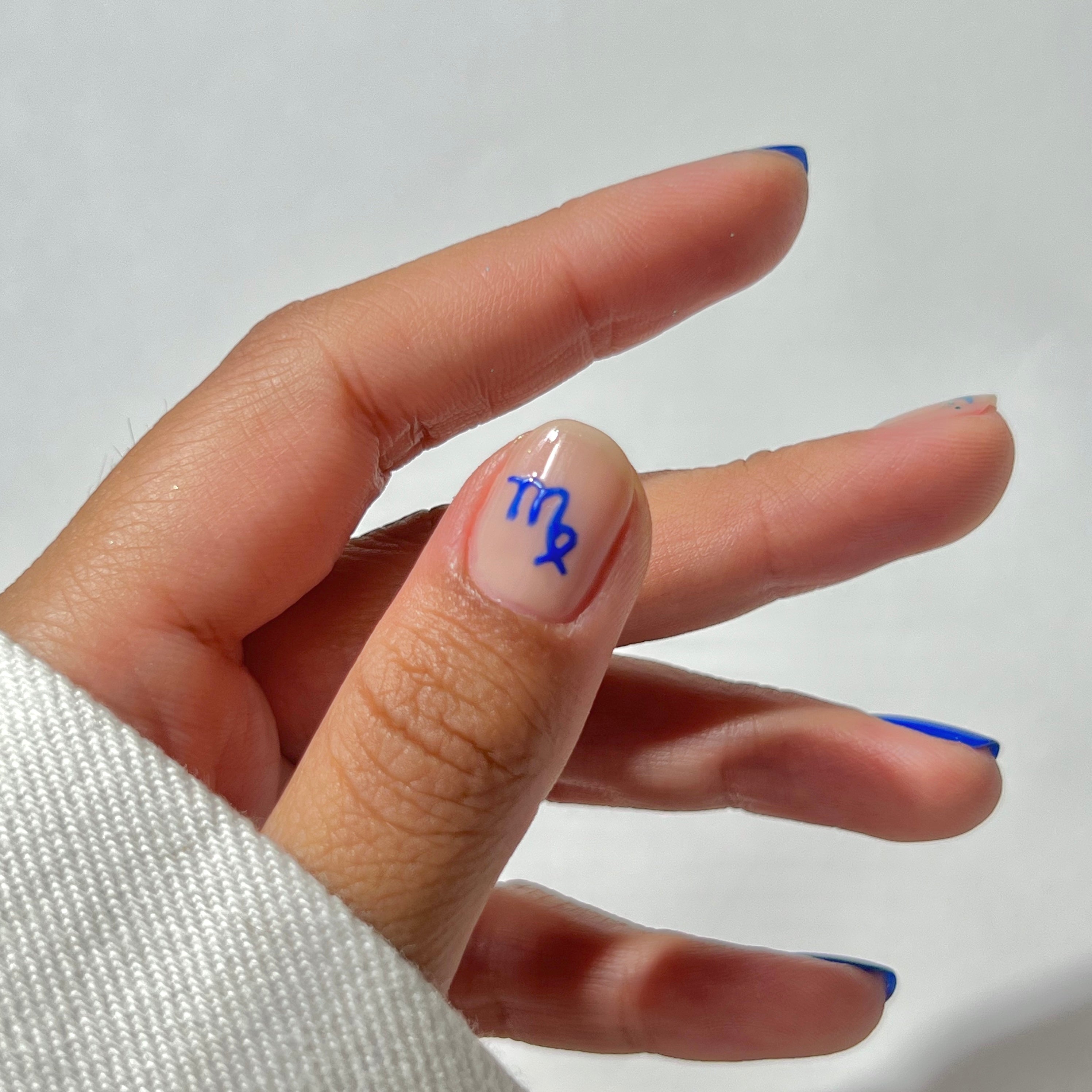 A hand with neatly manicured nails, featuring a blue Virgo zodiac symbol on the thumb. The nails have a natural base color with thin blue tips. The hand is partially covered by a white sleeve against a plain background.