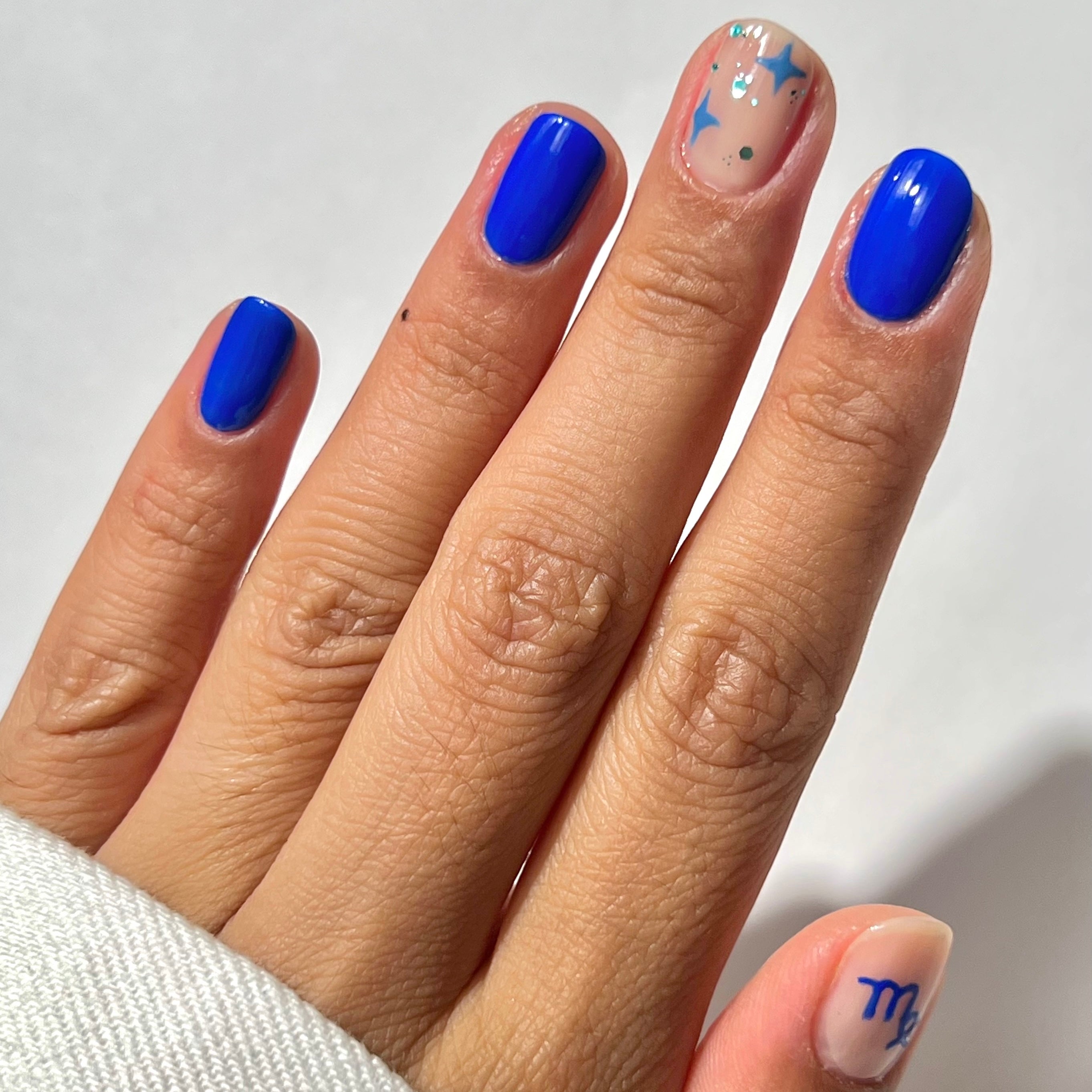 A hand with blue-painted nails; the ring finger has a nude base with blue and silver abstract shapes, and the pinky has a nude base with a blue Virgo zodiac symbol.
