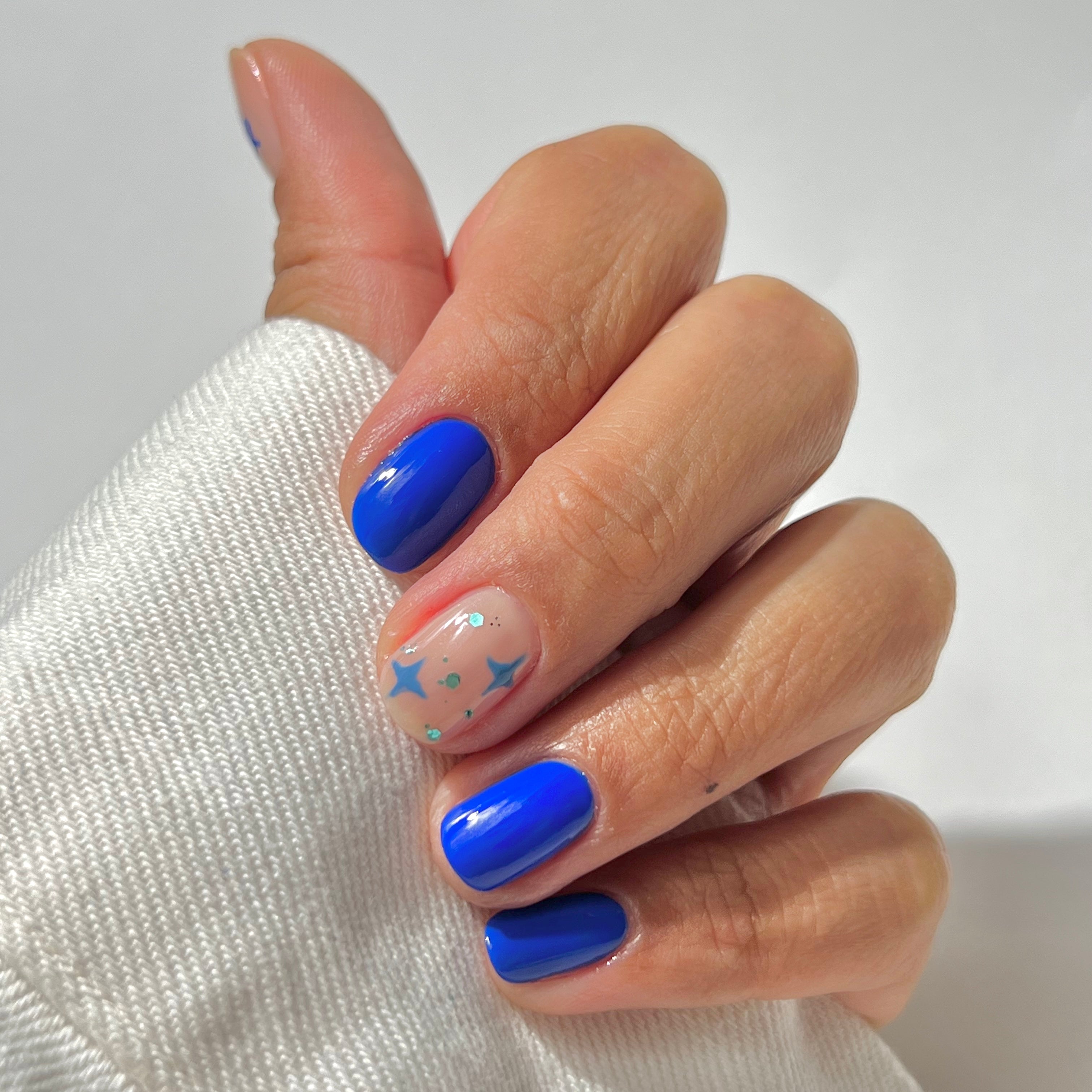 A hand with short, neatly manicured nails; four nails are painted bright blue, and the ring finger has a clear base with blue stars and small colorful dots as nail art. The person is wearing a white long-sleeve top.