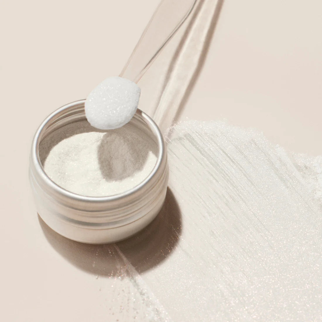 Un petit pot de poudre effet glazed blanc de Manucurist est ouvert sur une surface claire, avec une petite cuillère transparente tenant une dose au-dessus et une traînée de poudre à côté — idéal pour créer de superbes ongles glazed blancs lors de votre prochaine manucure.