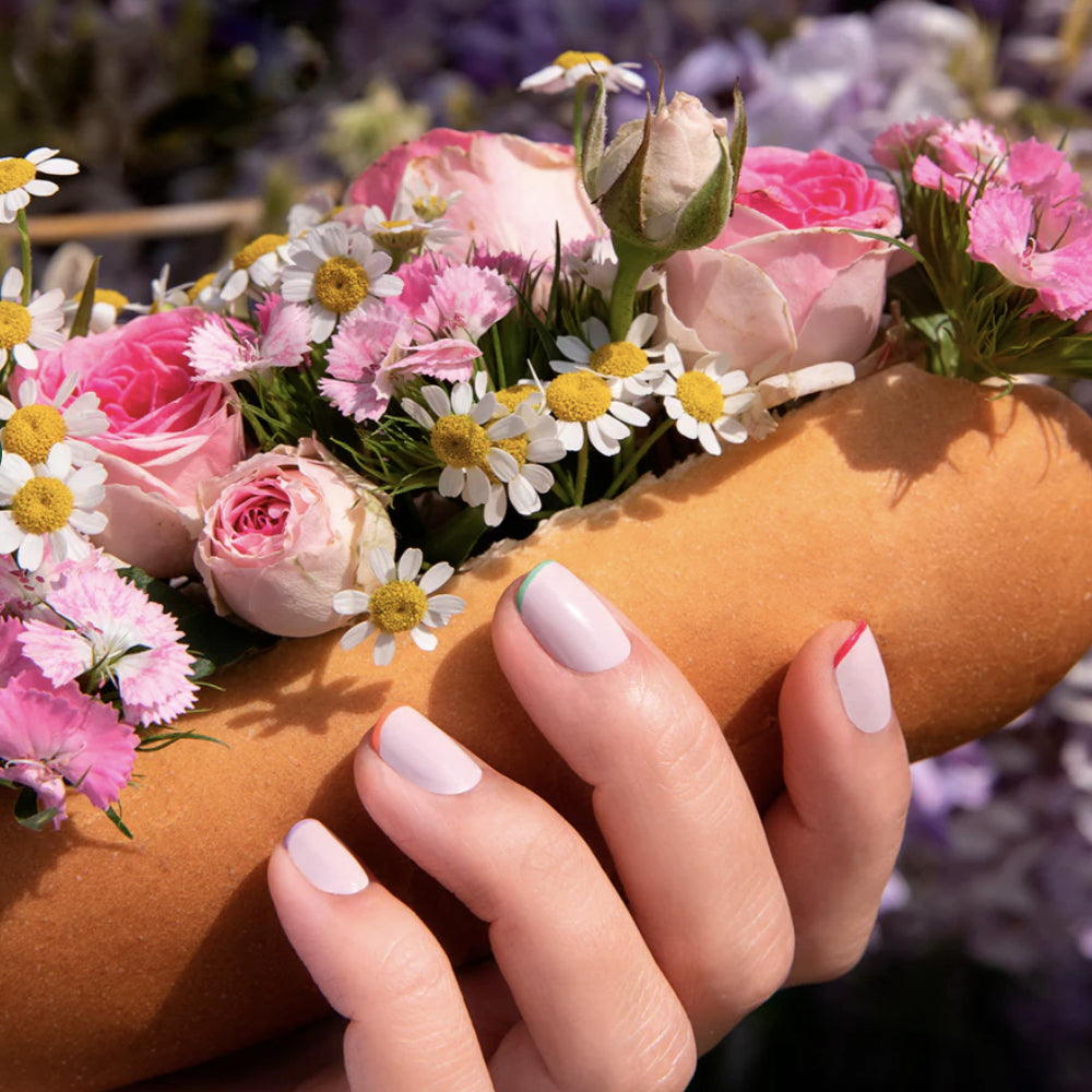 A hand with manicured nails holds a hot dog bun filled with pink roses, daisies, and other flowers instead of food, against a blurred floral background.