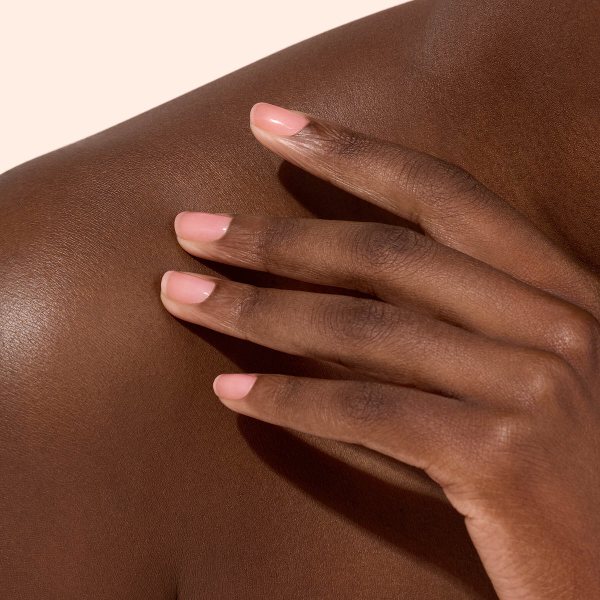 Gros plan sur une main aux ongles soigneusement manucurés, vernis avec Manucurist Active™ Glow Cranberry, dont le vernis éclatant brille sur une peau brun foncé lisse, posée délicatement sur une épaule nue, le tout sur un fond beige clair.