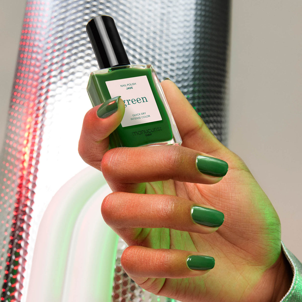 A hand with neatly manicured nails painted in "Jade" by Manucurist, a mineral green shade with cool undertones, holds the matching polish bottle against a metallic textured background with green and red light reflections.