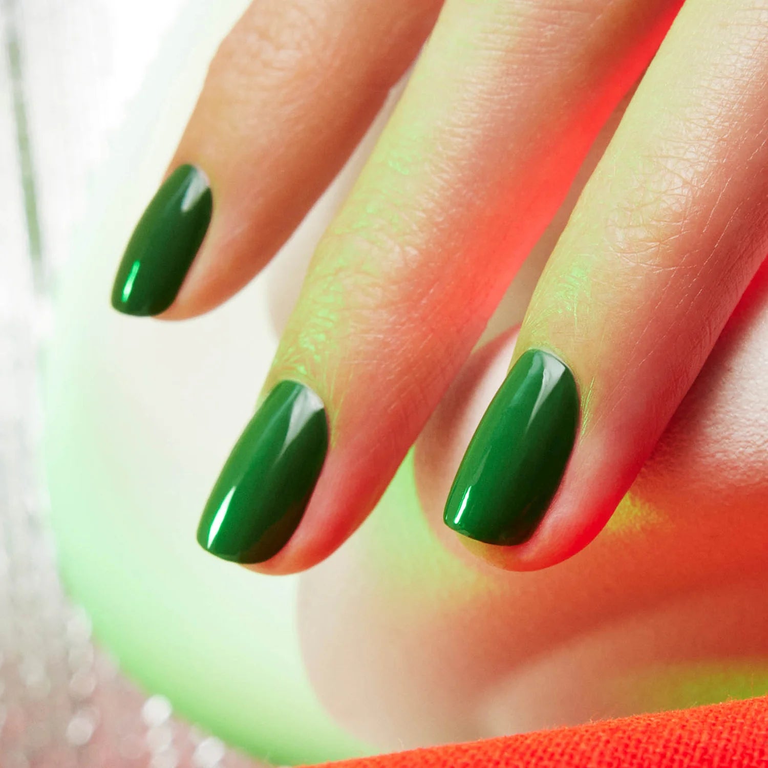 A close-up of a hand with shiny, dark green oval gel nails. The nails have a glossy finish and the background features soft red and green lighting.