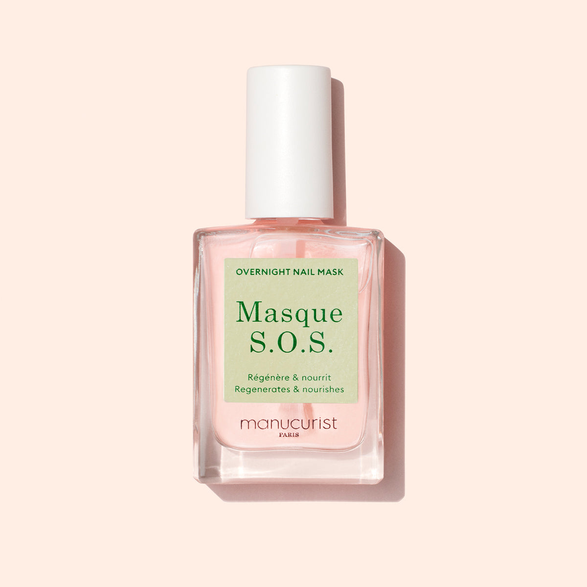 Un flacon de Manucurist S.O.S. Nail Mask, un soin hydratant pour les ongles avec un liquide rose et un bouchon blanc, est posé sur un fond rose clair. L’étiquette présente du texte en français et en anglais.