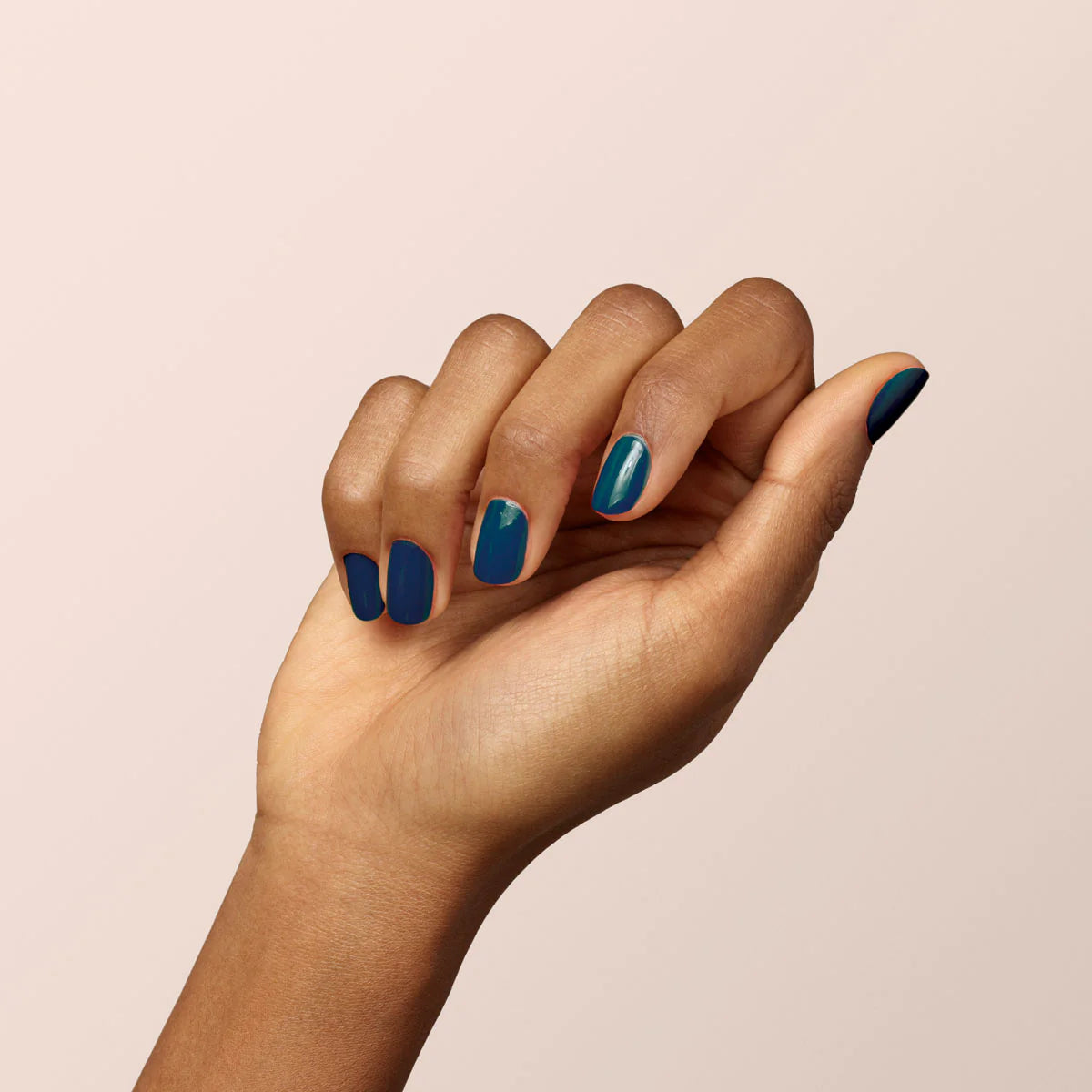 A hand with medium brown skin is held up against a light beige background. The nails are painted dark blue, with one nail featuring a small light blue and green accent design.