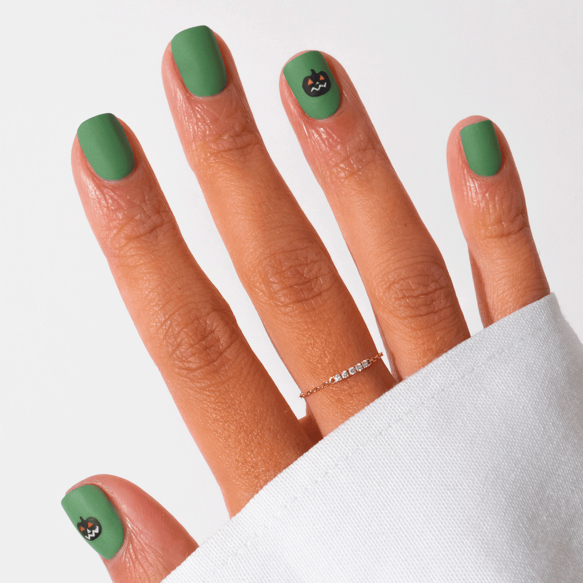 Une main avec un vernis gel vert mat Pumpkin Patch de Manucurist, orné de petits visages de chats noirs sur l’auriculaire et le pouce. Le look est complété par une fine bague dorée et une chemise blanche à manches longues — un nail art d’Halloween subtil et parfait.