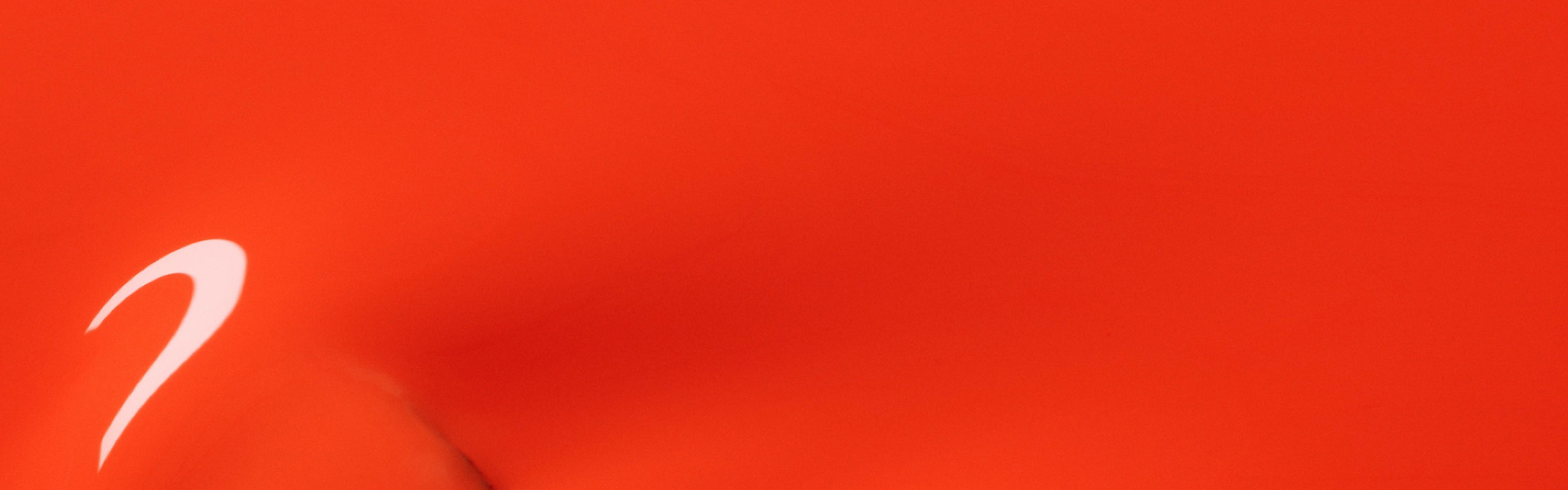 Close-up of a smooth, bright orange surface with a small, white curved mark or reflection on the left side. The image has an abstract and minimalistic appearance.
