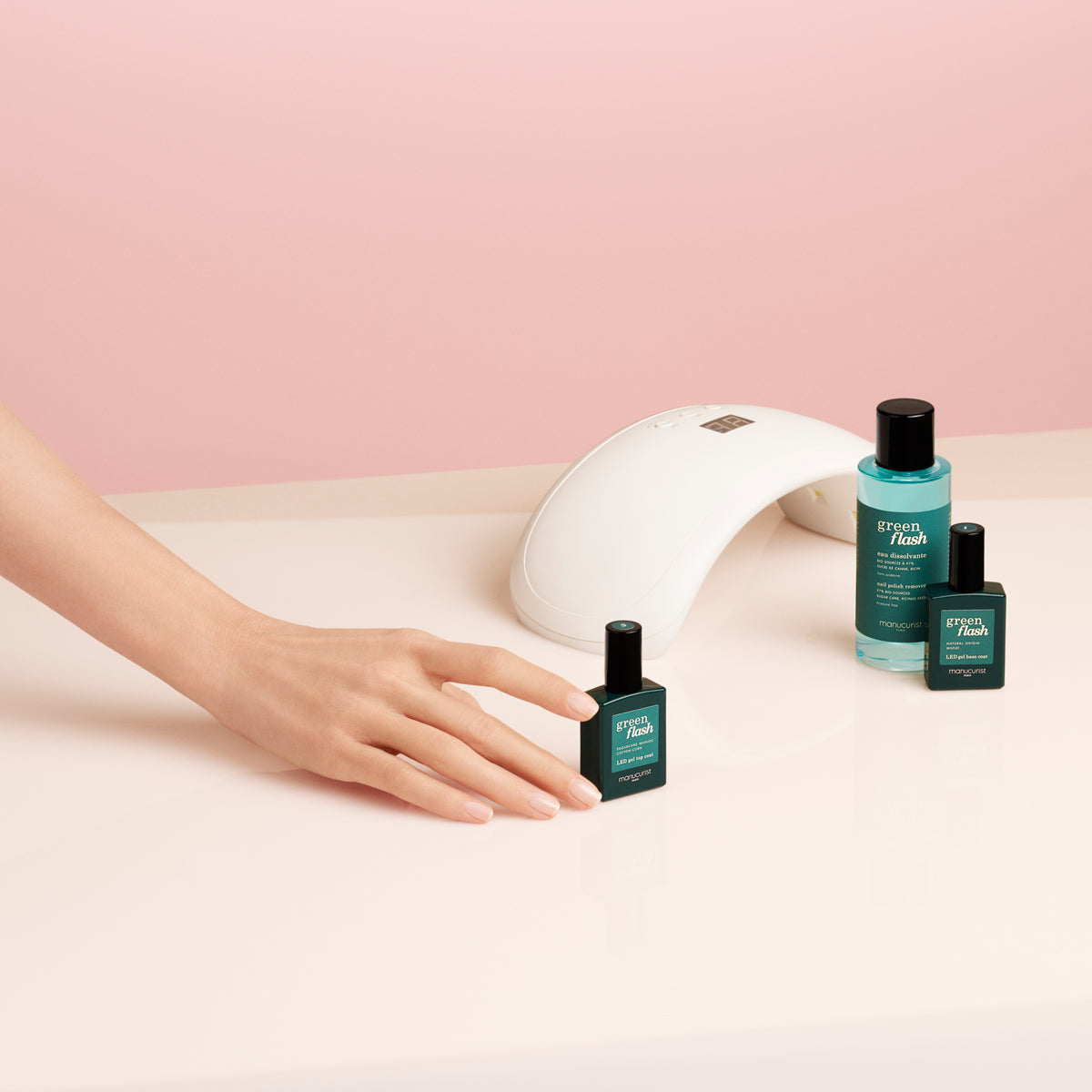 A hand reaches for a Manucurist-test Welcome kit Green Flash nail polish bottle on a light pink surface, with two other bottles and a white nail lamp nearby against a pink background.