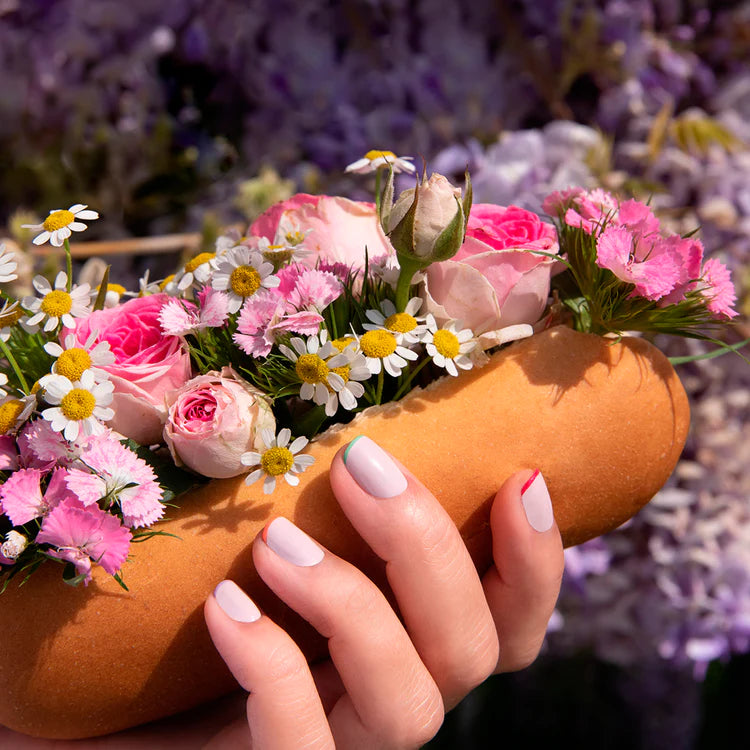 A hand with pastel-painted nails holds a hot dog bun filled with pink roses, daisies, and various small flowers, instead of food. The floral arrangement is vibrant, set against a blurred background of greenery and blossoms.