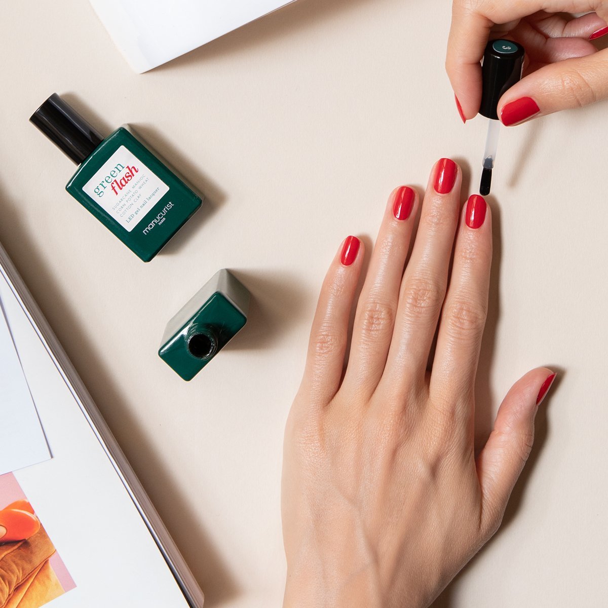 A person applies clear top coat from the Manucurist-test Welcome kit Green Flash to their red-painted nails. Two bottles of nail polish and an open magazine rest on a beige surface beside their hand.