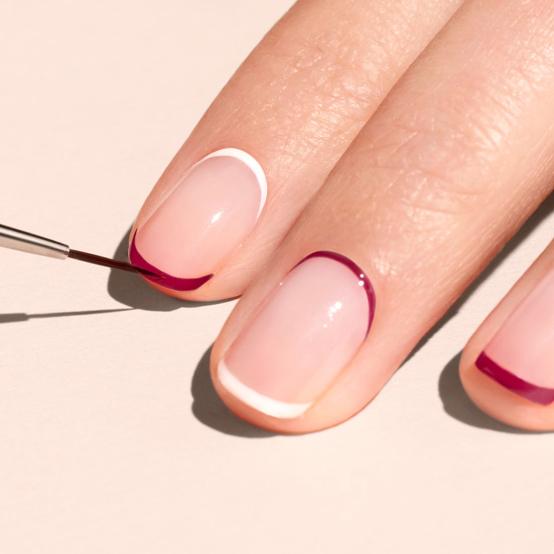 A hand with neatly manicured nails shows minimalist nail art; the Manucurist Liner Brush uses vegan synthetic bristles to paint deep pink curves on a neutral base, delivering professional-quality details and a crisp white-tipped nail.