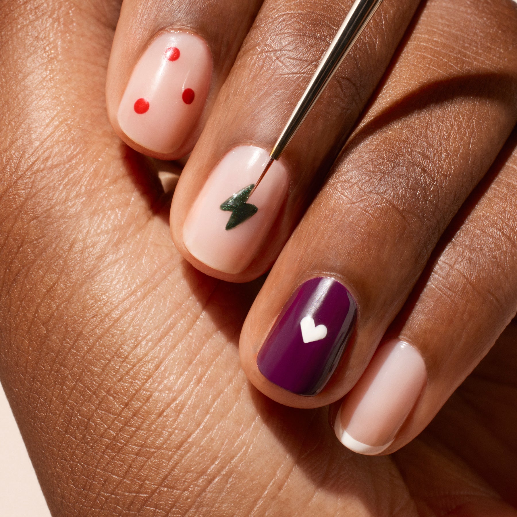 A manicured hand displays nail art—red dots, a green lightning bolt, a purple nail with a white heart, and natural nails with white tips and hearts—as the Manucurist Fine Tip Brush with vegan synthetic bristles touches one nail.