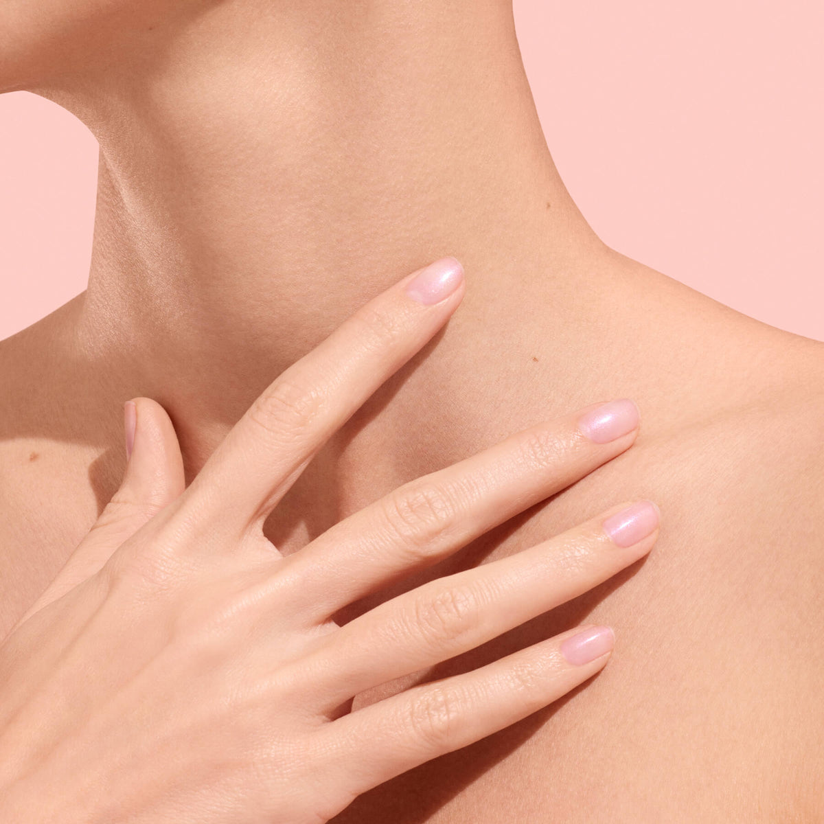 Gros plan sur la main d’une personne — présentant des ongles lisses et hydratés grâce au soin des ongles végétal Active™ Plump Pink Glazed de Manucurist — touchant délicatement son cou et sa clavicule, avec une peau lisse et un fond rose clair.