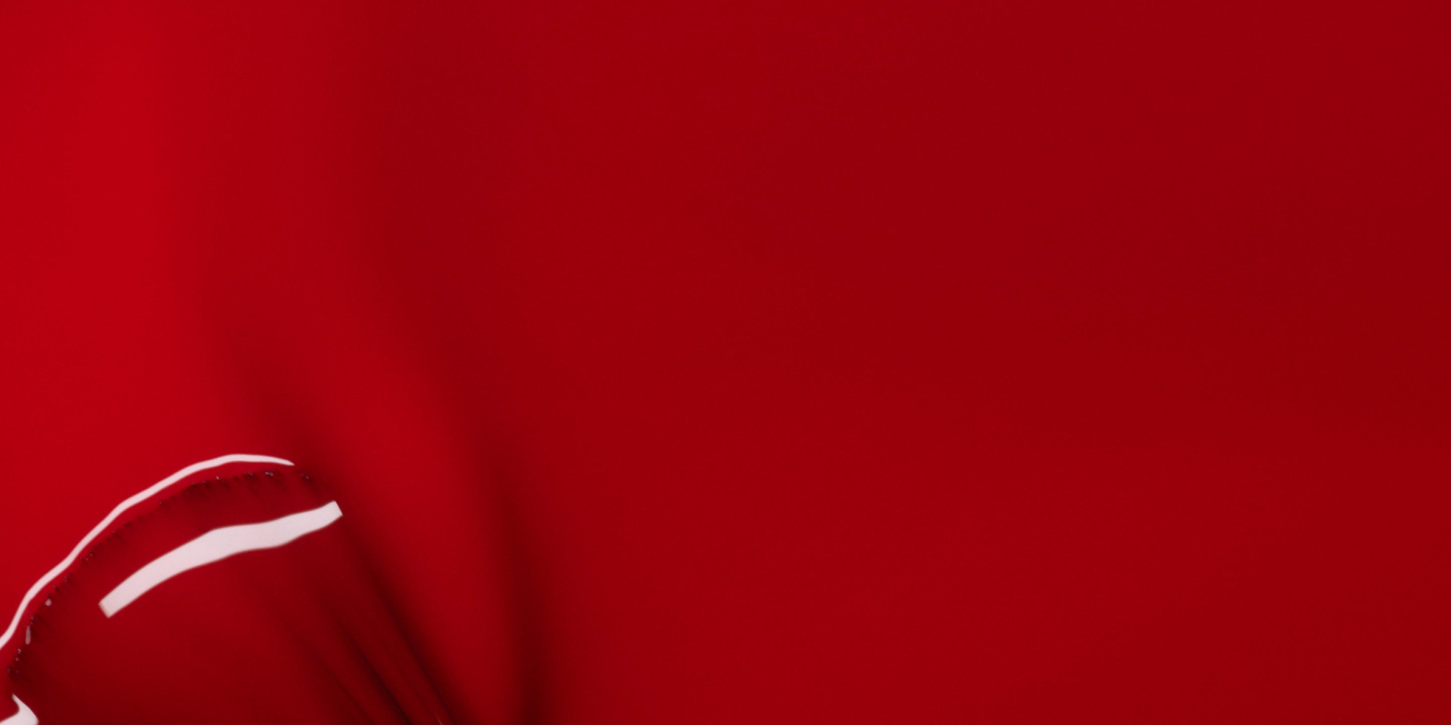 Close-up of a smooth, red surface with a glossy shine and a small white highlight near the bottom left corner, possibly from a curved or folded material. The background is a consistent, vibrant red.