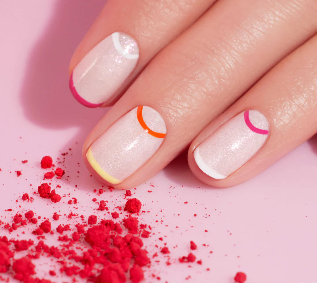 A hand with short, neatly manicured nails painted in pale pink, each tipped with a colorful, curved line in pink, orange, purple, or yellow. Red powder is scattered on the pink surface beside the fingers.