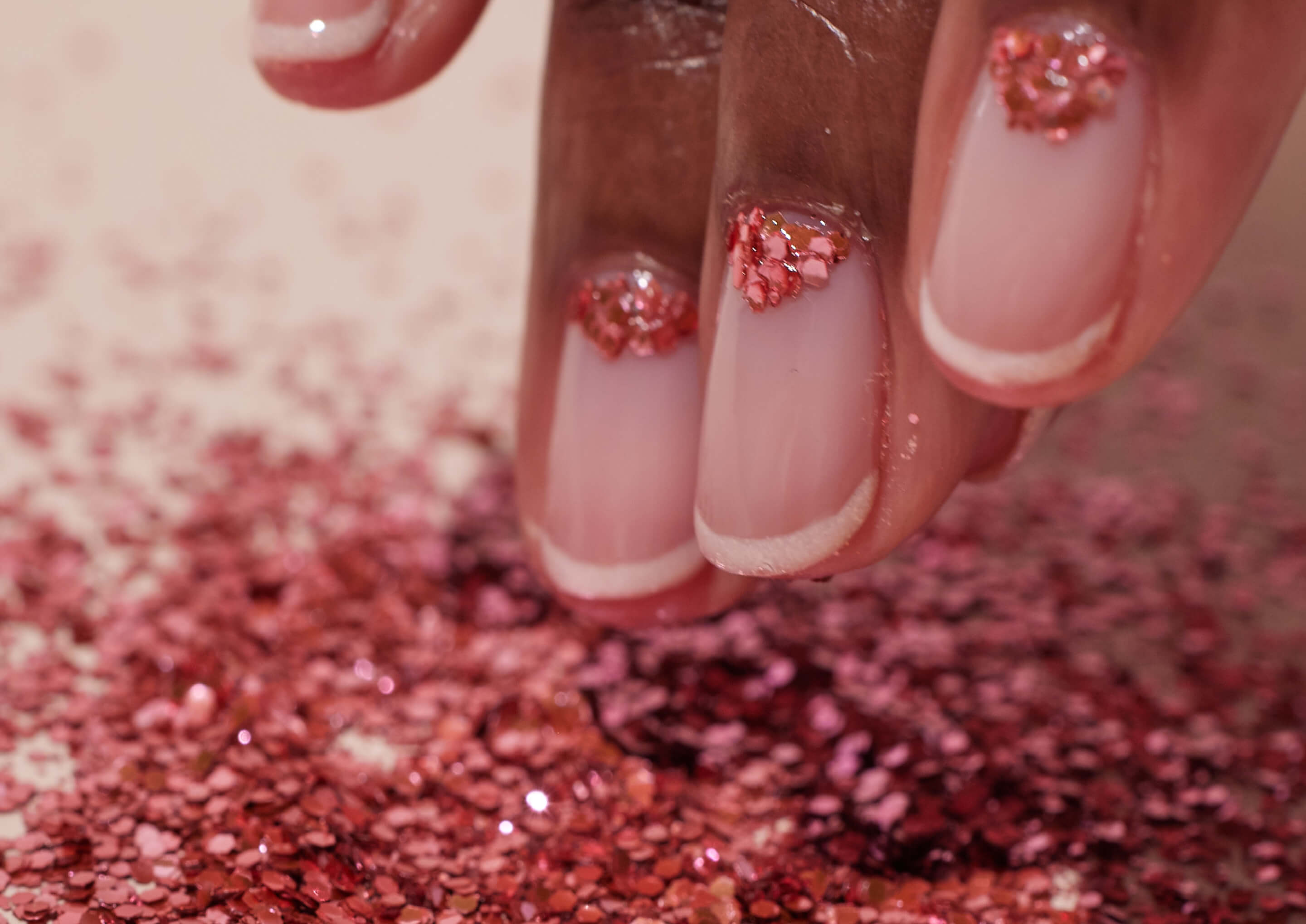 A close-up of a hand with neatly manicured nails, featuring nude polish and pink glitter accents, gently touching a surface covered in sparkling pink glitter.