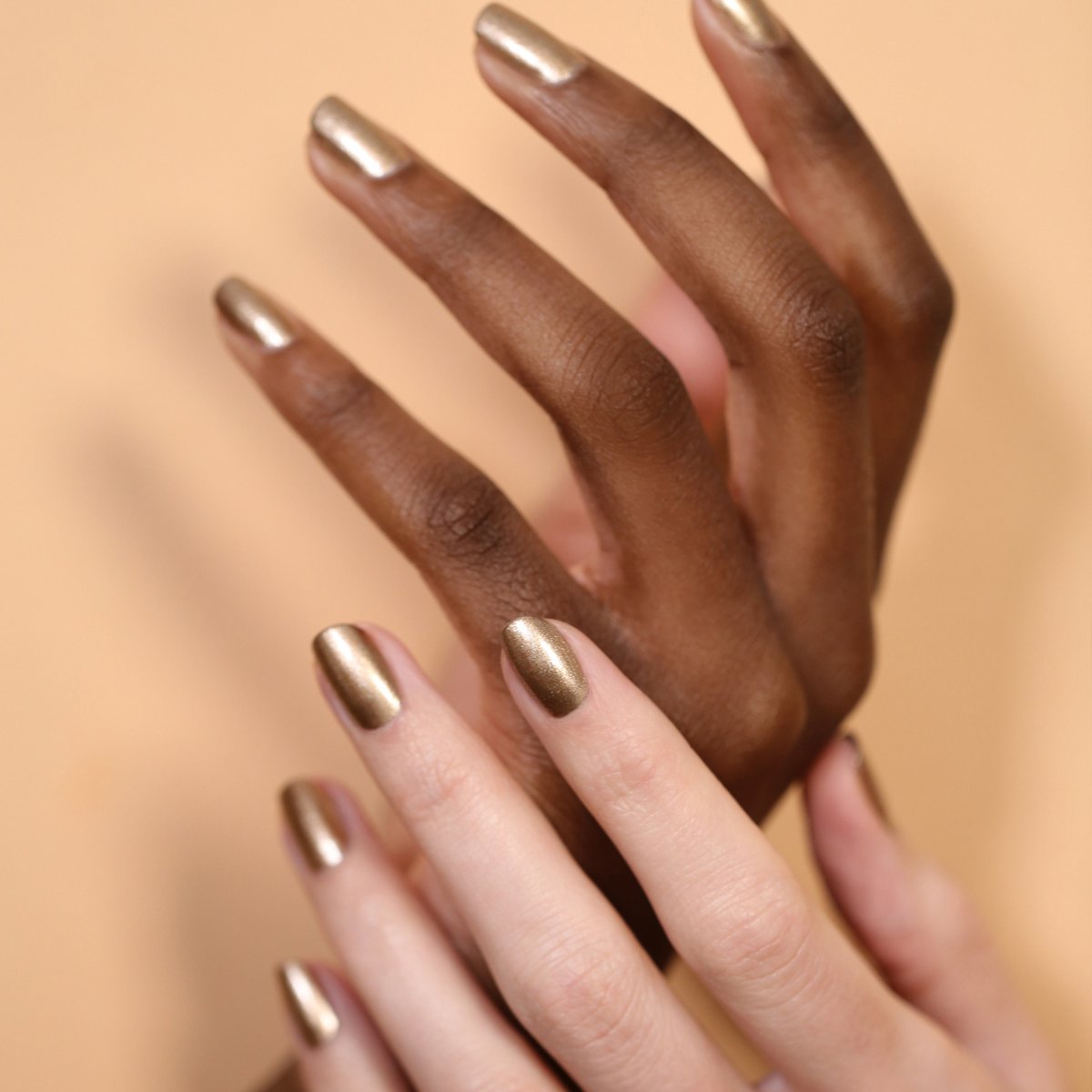 Deux mains aux carnations différentes, chacune arborant des ongles courts et manucurés vernis avec le gel métallique Bronzé de Manucurist, joliment mises en scène sur un fond beige doux.