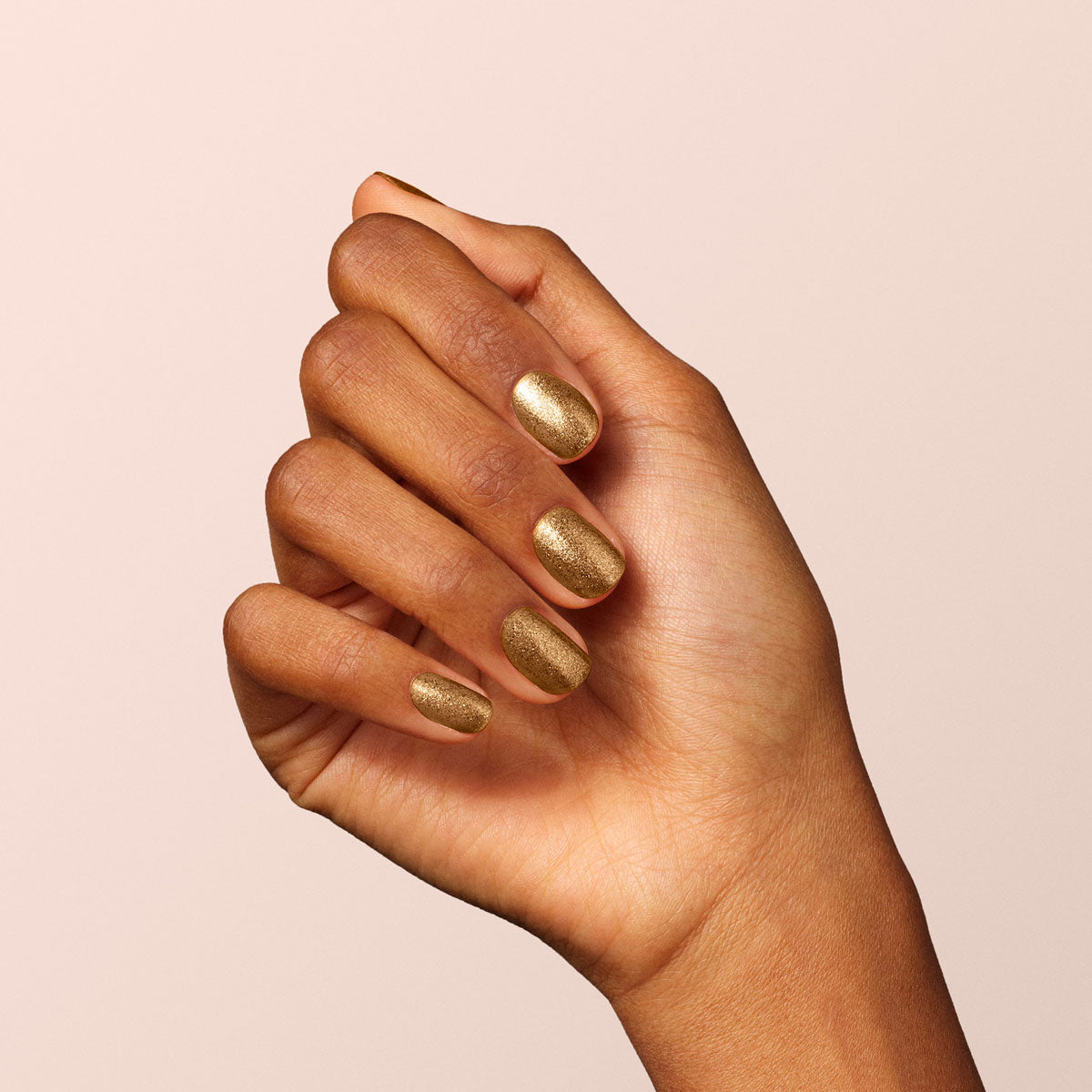 Une main à la peau brun moyen, les doigts légèrement recourbés vers l’intérieur, présente des ongles peints en Bronzé de Manucurist — un vernis gel doré et cuivré scintillant — sur un fond beige clair.
