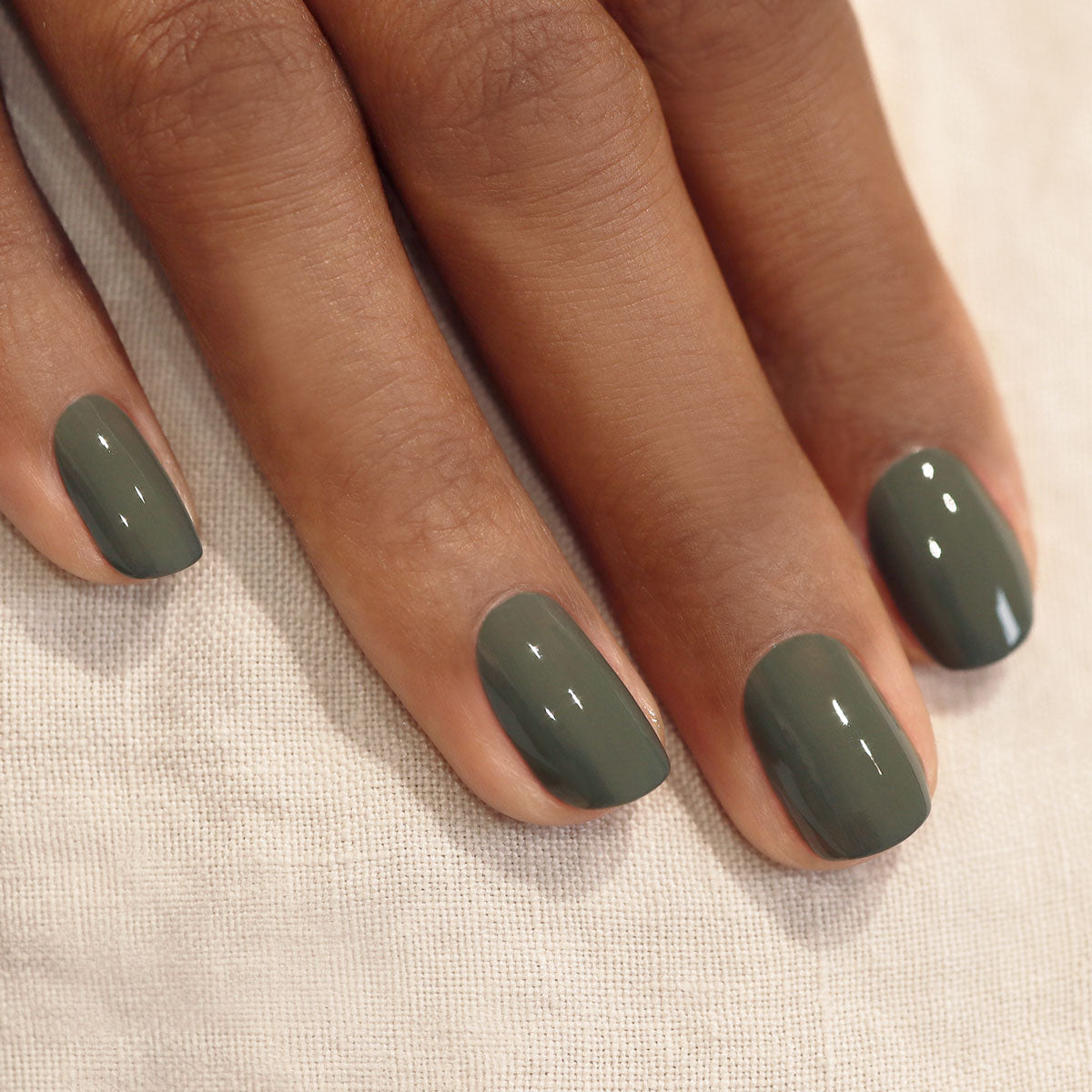 Gros plan d’une main aux ongles parfaitement manucurés, peints avec le vernis Khaki brillant de Manucurist, reposant sur un tissu clair texturé.