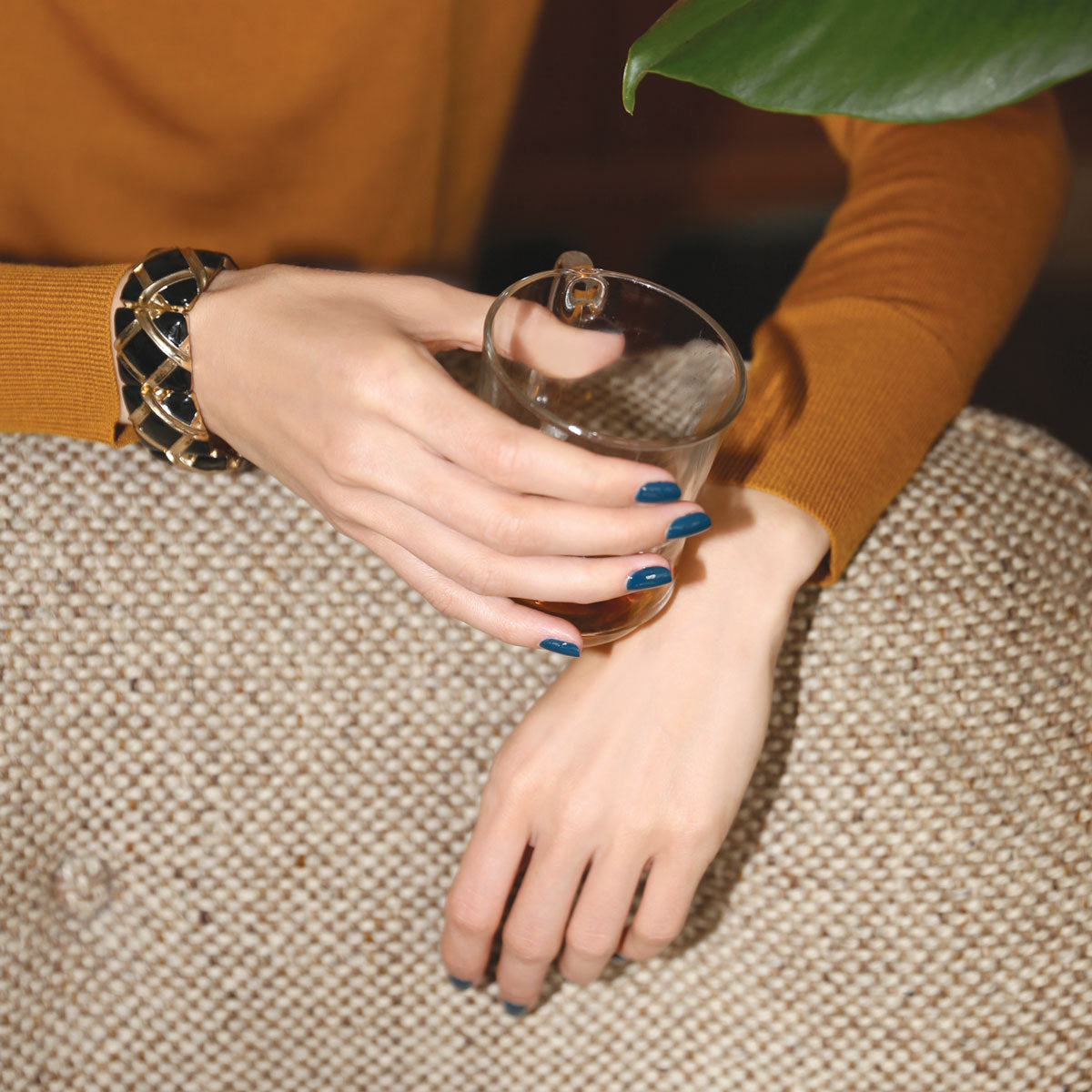 Wearing Dark Clover by Manucurist, a person with teal nails holds a glass of brown liquid, dressed in a mustard top with a black and gold bracelet, relaxing on a textured beige surface beside a green leaf.