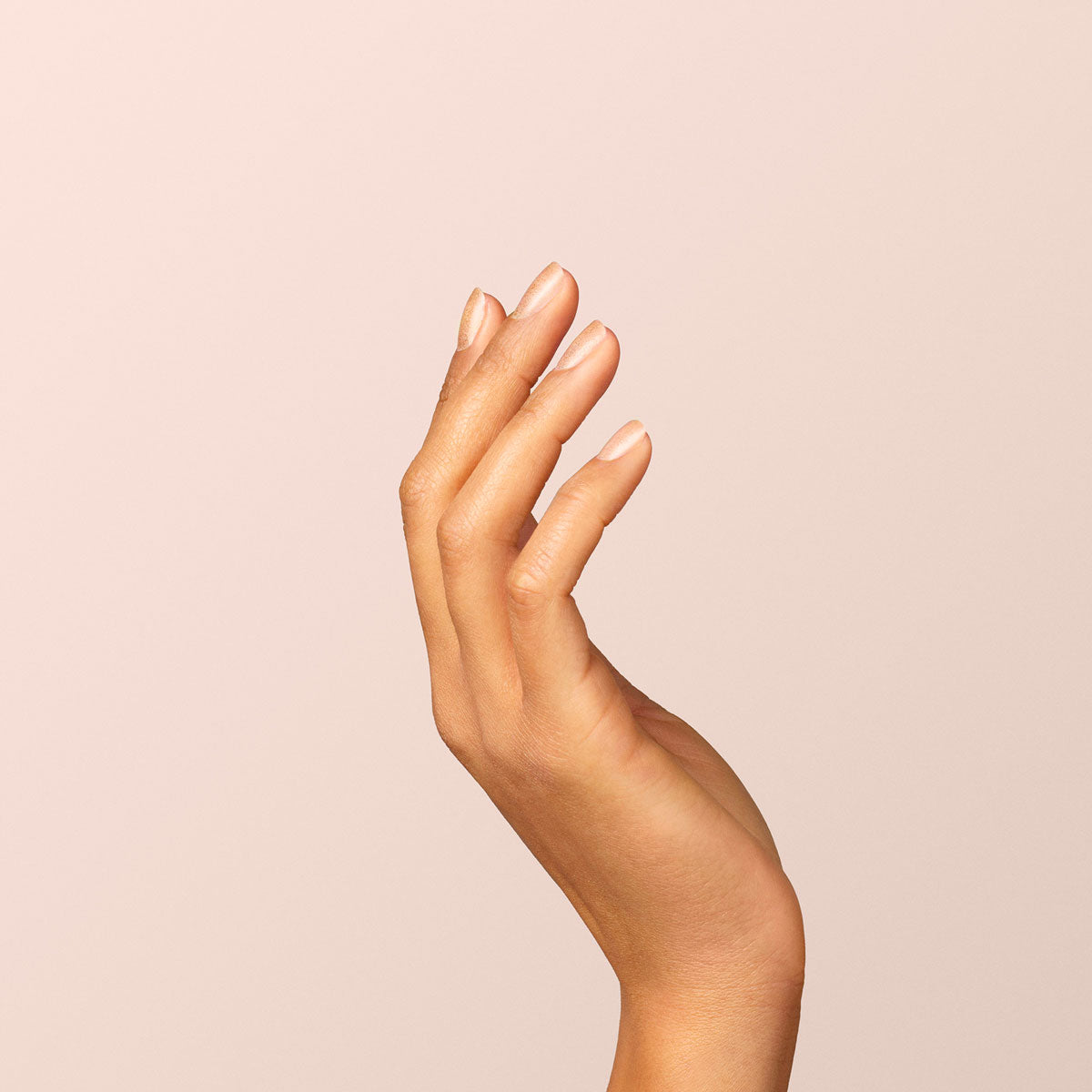 A hand with neatly manicured nails, some coated in Manucurist’s Gold polish, is raised against a plain light beige background. The fingers are slightly curved and relaxed, emphasizing smooth skin and elegant, natural nails.